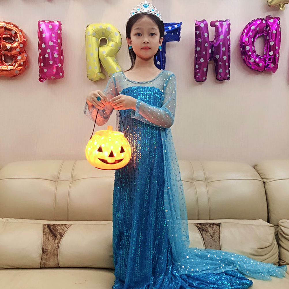 Ein Mädchen in blauem Kleid und Krone hält eine Kürbislaterne. Ballons schreiben 'BIRTHDAY' an die Wand. Im Hintergrund ein beiges Sofa.