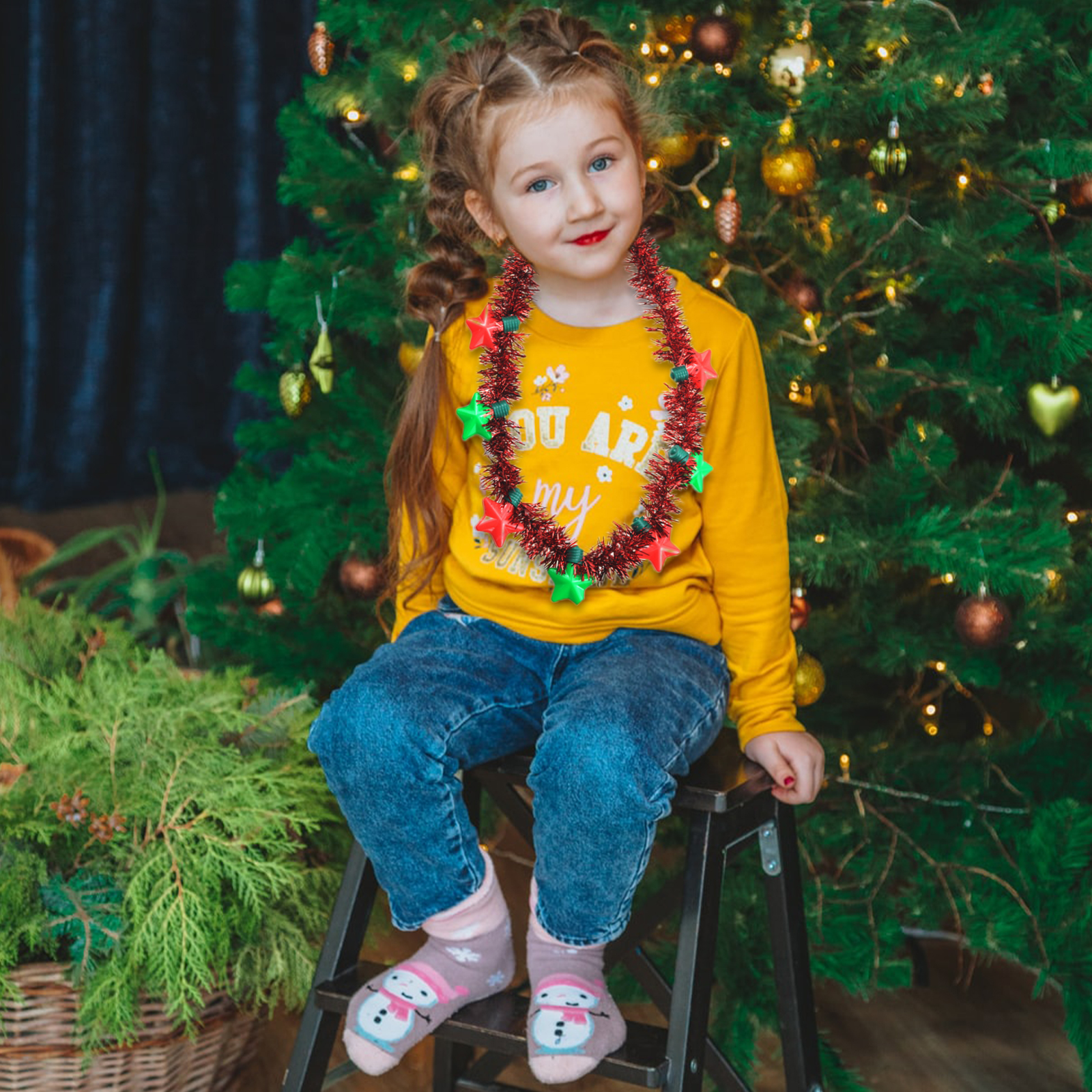 Mädchen in gelbem Pullover mit Sternengirlande sitzt vor einem Weihnachtsbaum. Sie trägt Jeans und Schneemannsocken.