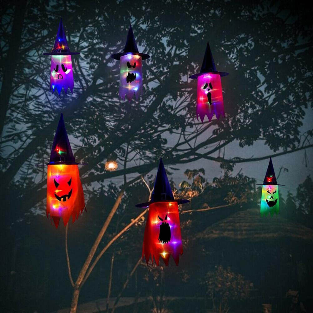 Halloween-Dekorationen: leuchtende geisterartige Laternen hängen an einem Baum. Jede hat einen Hexenhut und ein gruseliges Gesicht.