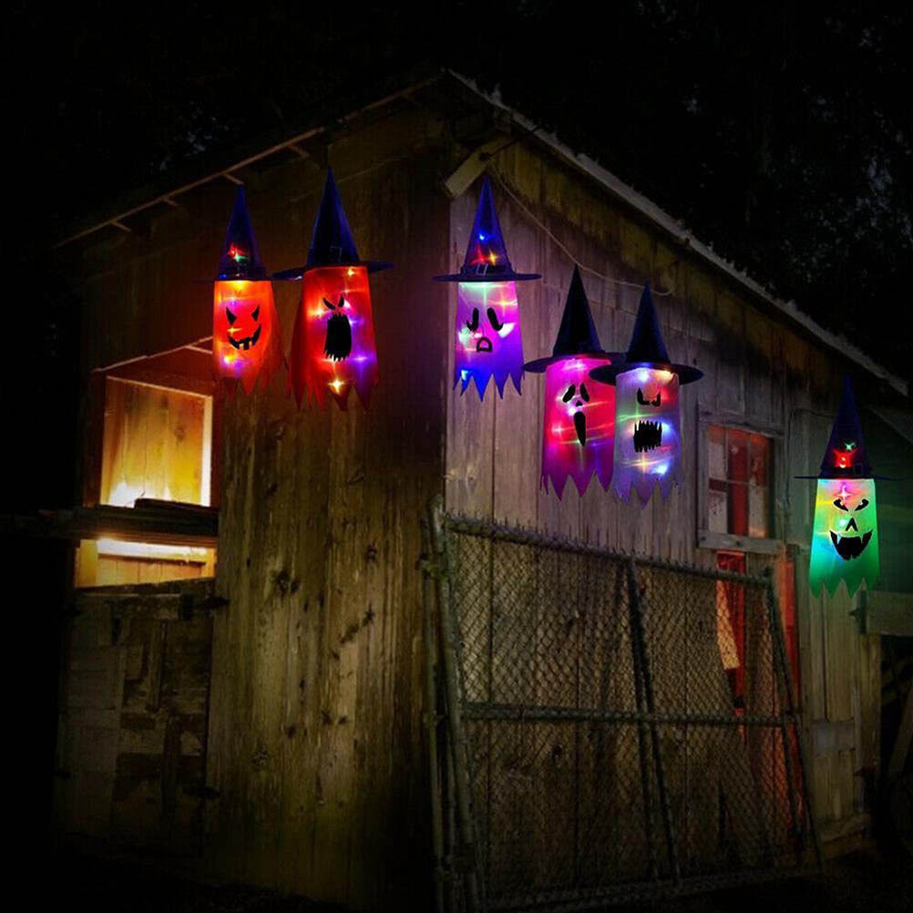 Halloween-Dekorationen: Leuchtende Geisterfiguren mit Hexenhüten hängen nachts vor einem Holzhaus.