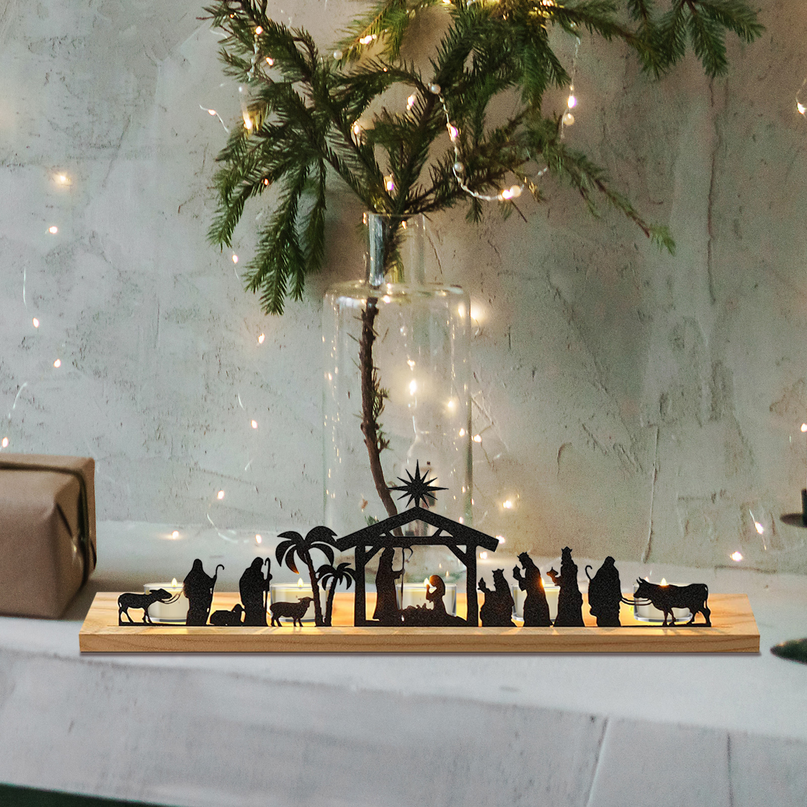 Eine Krippensilhouette auf einem Holzsockel mit Kerzen. Grün in einer Glasvase, Lichterketten und ein Geschenk sind sichtbar.