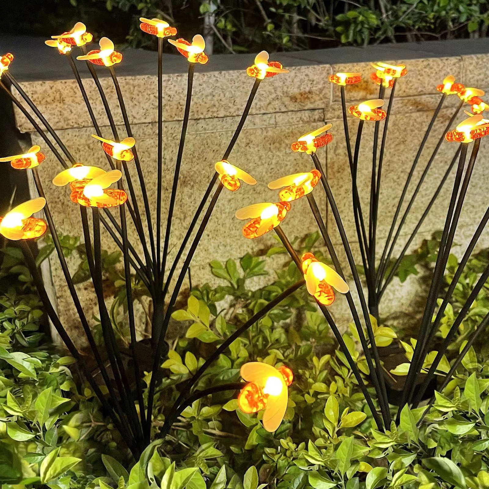 Dekorative Lichter in Form von Bienen mit orangefarbenen Flügeln und gelben Lichtern auf schwarzen Stielen, inmitten grüner Pflanzen.