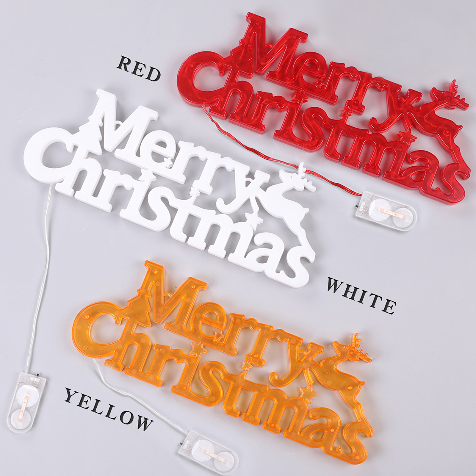 Drei Schilder mit der Aufschrift 'Merry Christmas' in Rot, Weiß und Gelb mit Rentieren, beleuchtet von kleinen Akkus, auf grauem Untergrund.