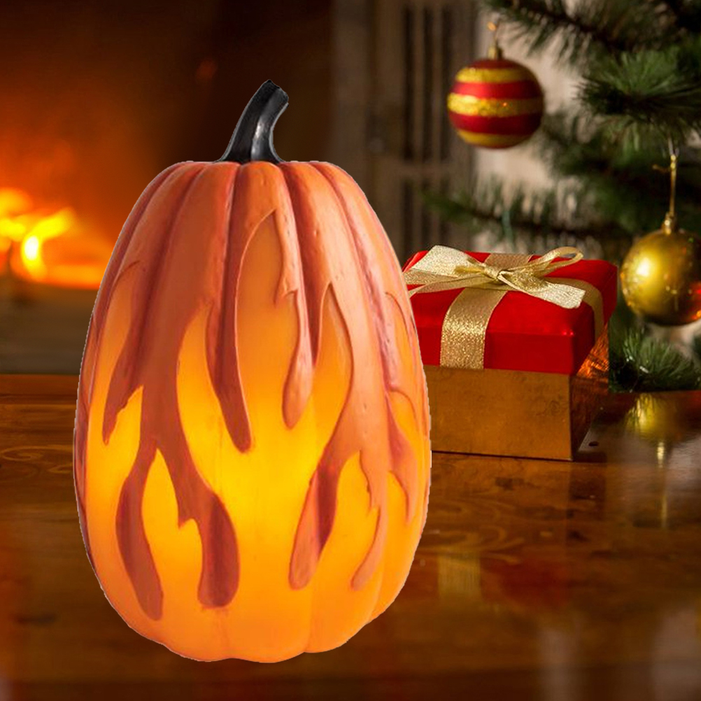 Eine kürbisförmige Lampe mit Flammendesign steht auf einem Tisch. Eine rote Geschenkbox mit goldenem Band und Christbaumschmuck sind zu sehen.