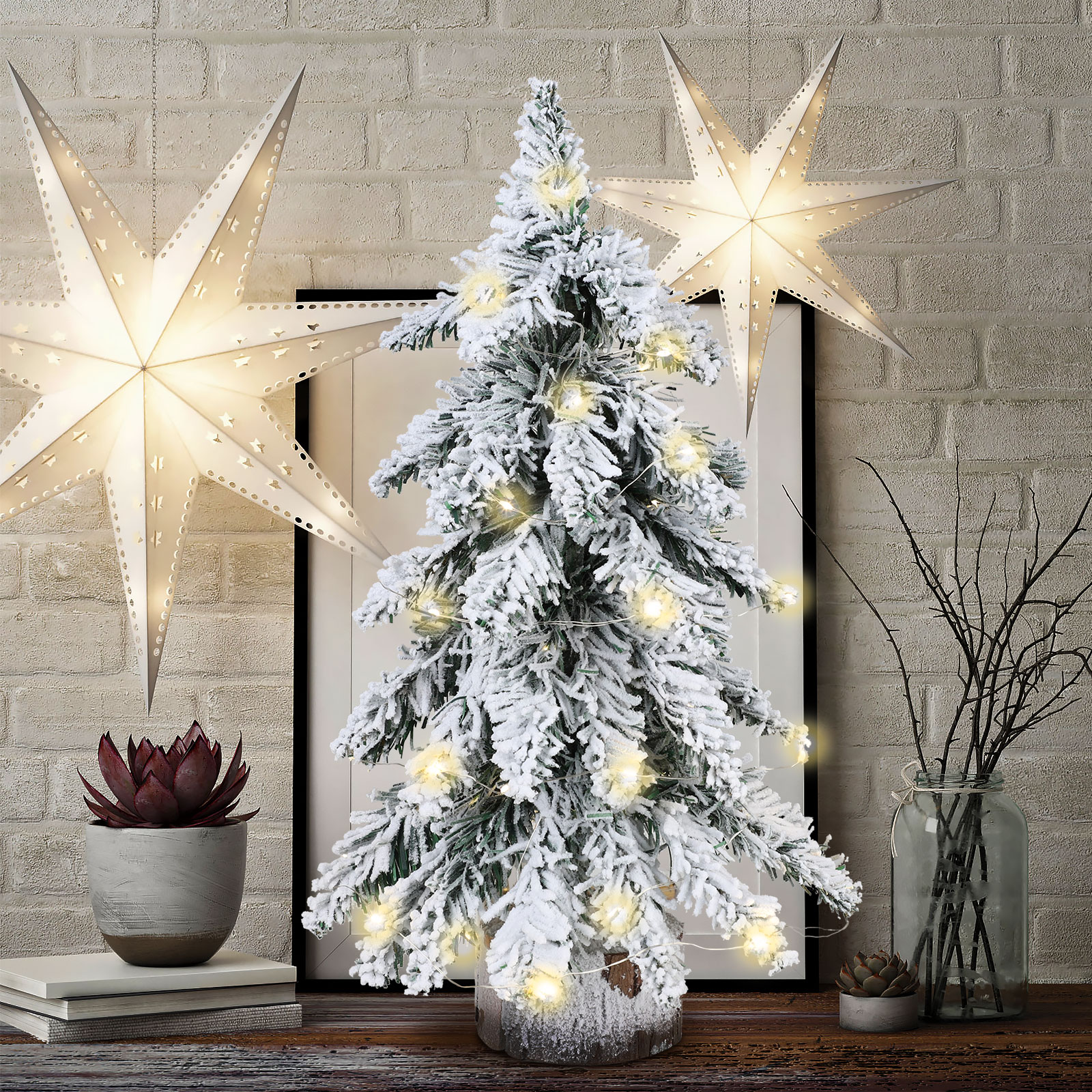 Ein kleiner, schneebedeckter Weihnachtsbaum mit Lichtern, zwei Sternen und einer roten Sukkulenten auf einem Holztisch.