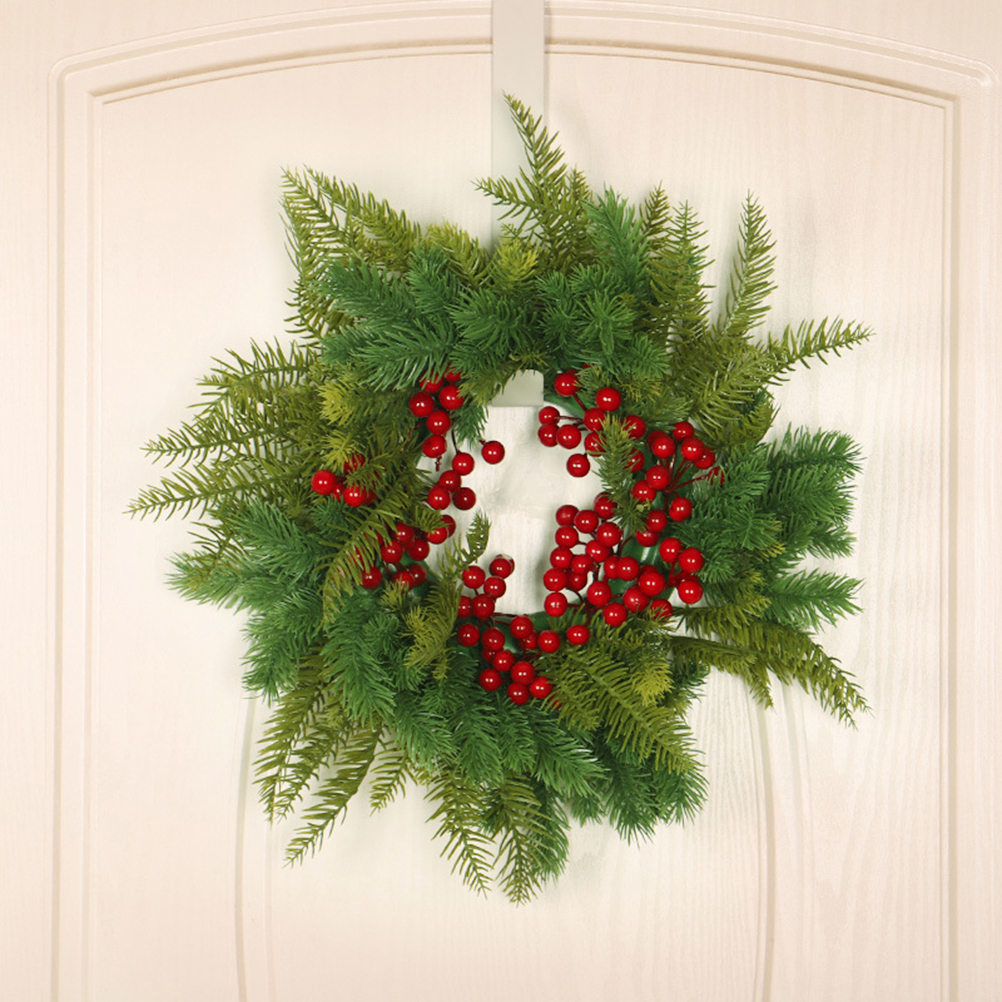 Ein Weihnachtskranz mit grünen Tannennadeln und roten Beeren hängt an einer weißen Tür. Er wird von einem weißen Türhänger gehalten.