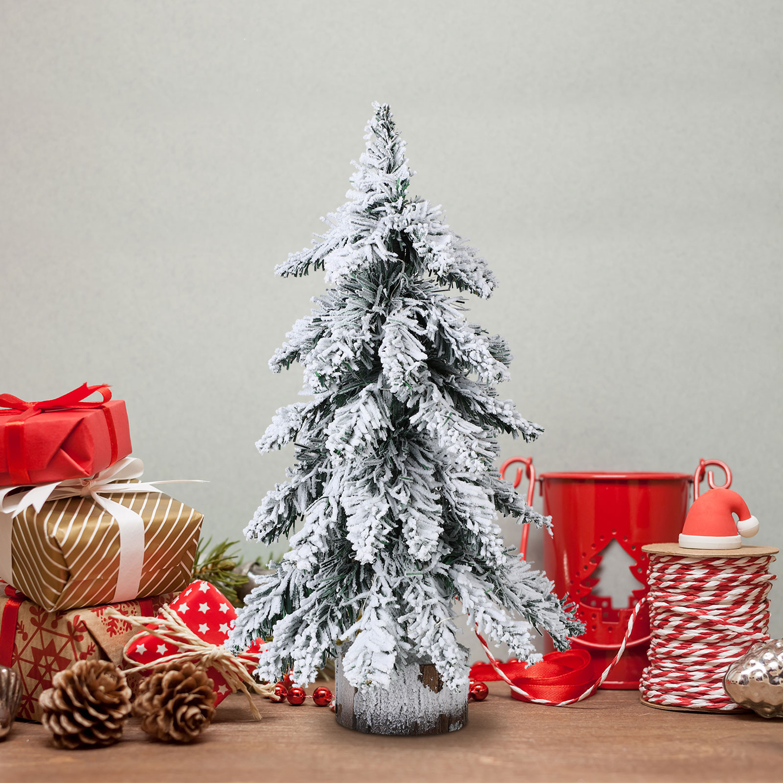 Ein verschneiter Weihnachtsbaum, Geschenke, Tannenzapfen, roter Eimer, Weihnachtsmütze und Dekorationen auf Holz.