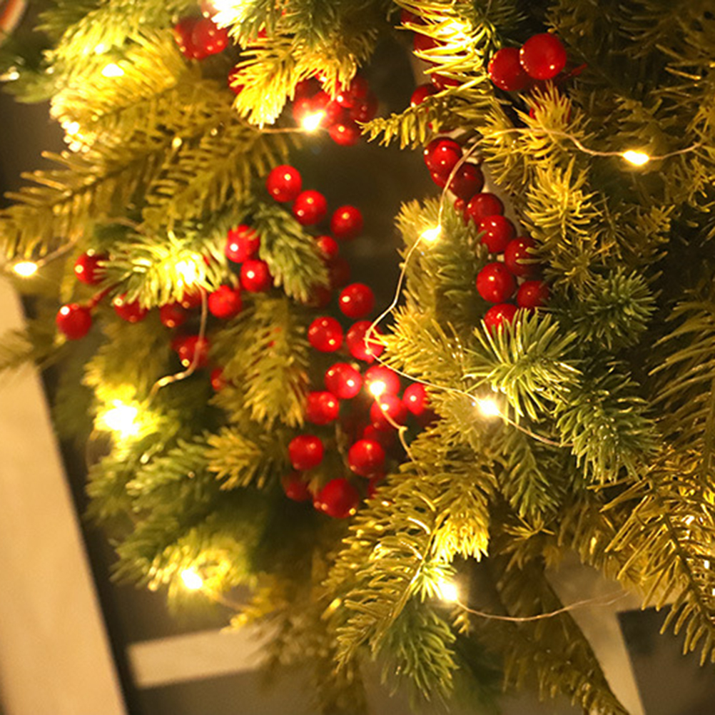 Nahaufnahme eines Weihnachtskranzes mit roten Beeren, grünen Tannennadeln und kleinen weißen Lichtern. Festlich und warm.