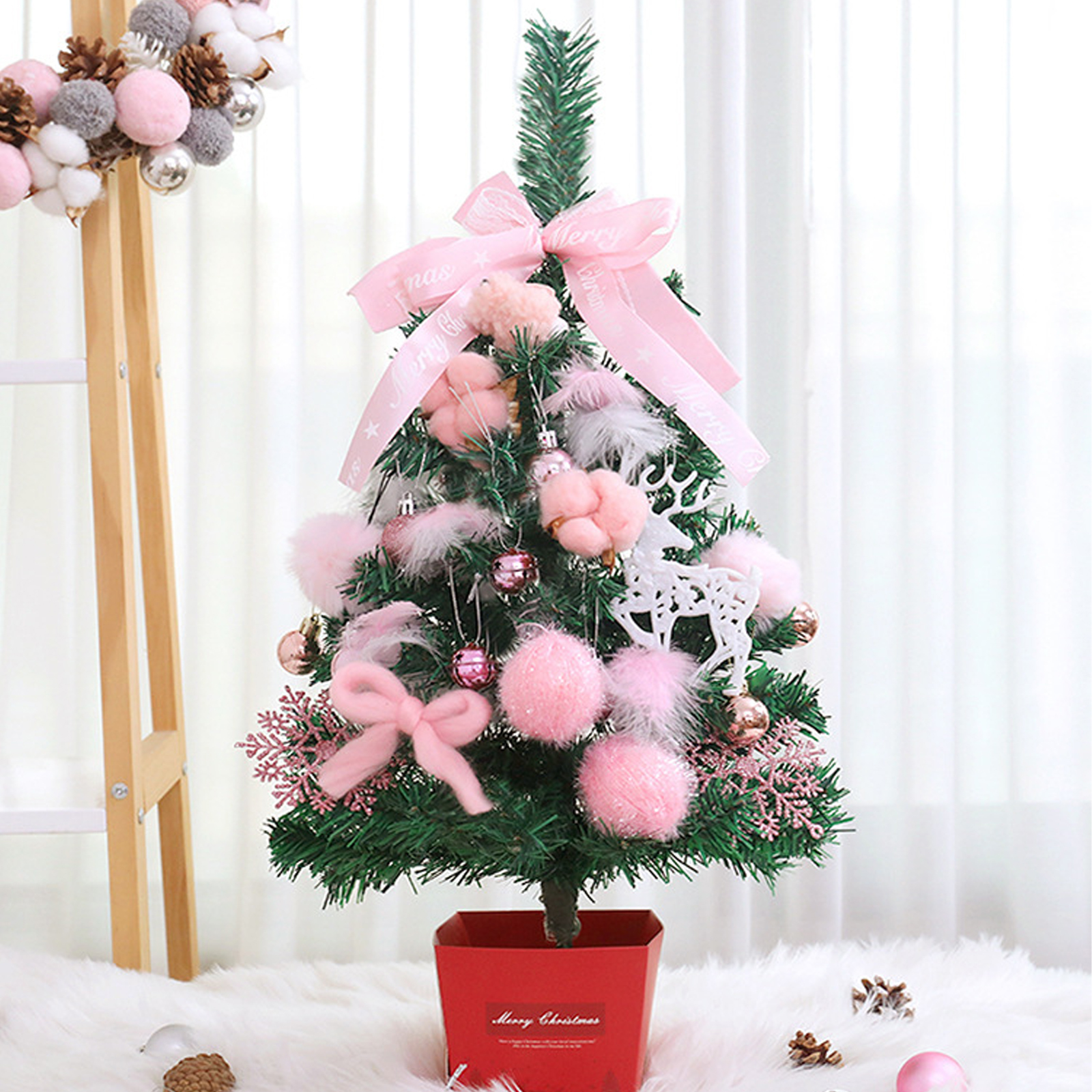 Ein kleiner, dekorierter Weihnachtsbaum in einem roten Topf. Er hat rosa Ornamente, eine Schleife und ein Band. Der Hintergrund ist ein weißer Vorhang.