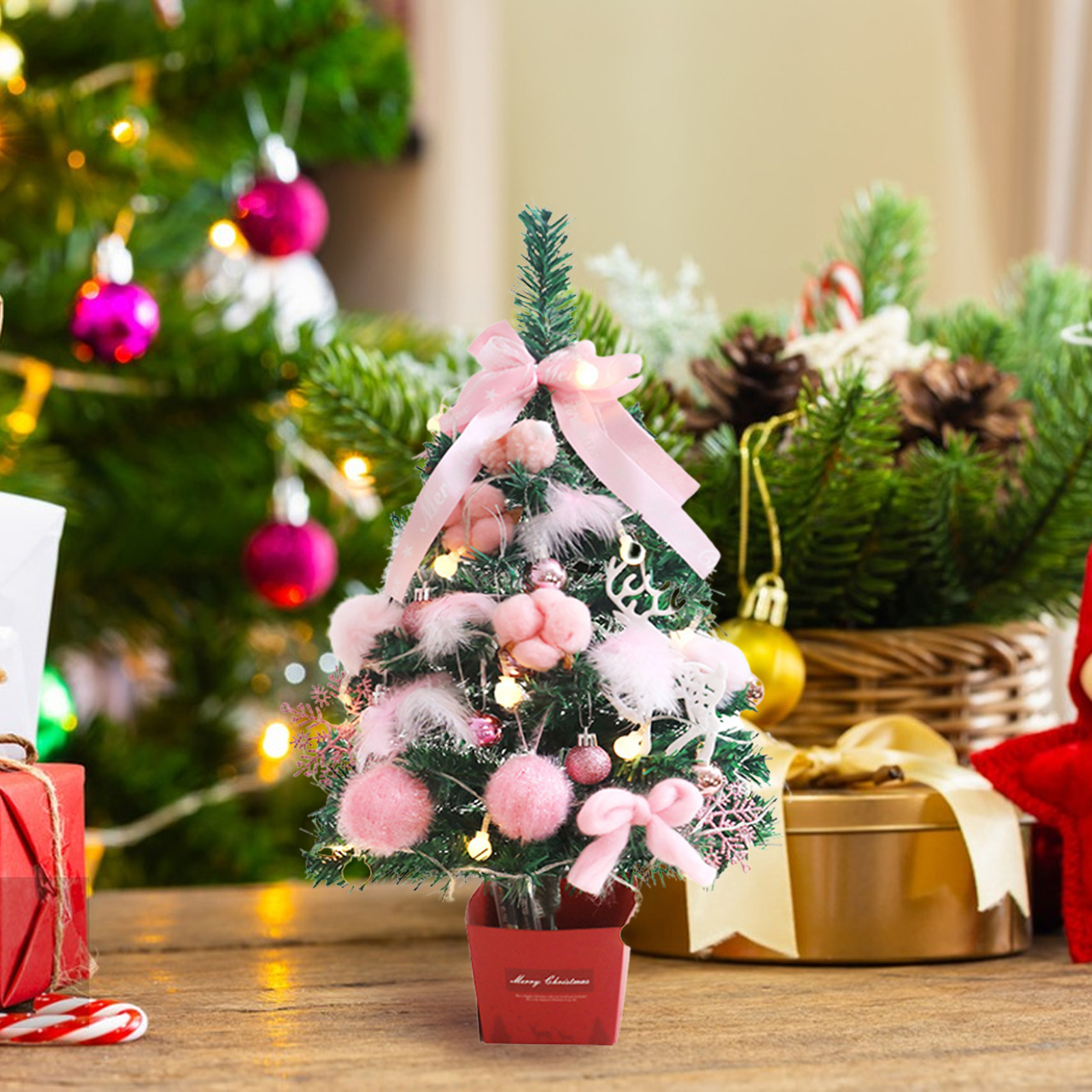 Ein kleiner, dekorierter Weihnachtsbaum mit rosa Ornamenten und Schleifen steht auf einer Holzoberfläche. Im Hintergrund Geschenke und andere Dekorationen.