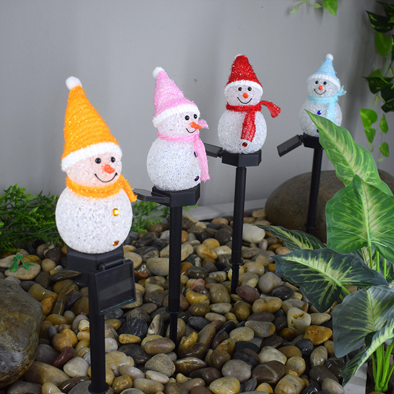 Vier Schneemann-Gartenleuchten mit unterschiedlich farbigen Hüten und Schals auf einem Kiesbett mit Grün.