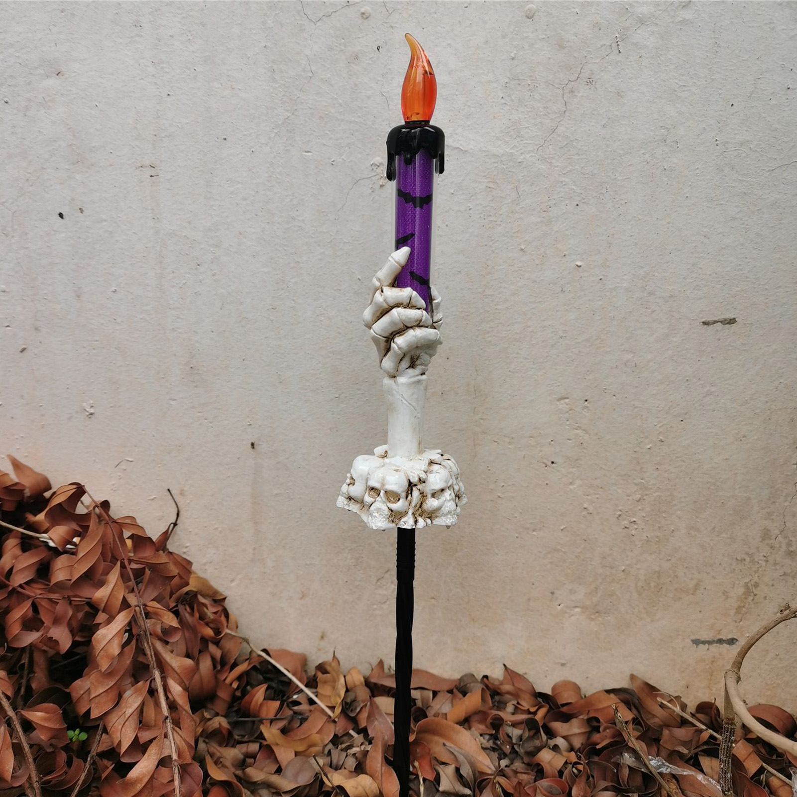 Eine Halloween-Kerzendekoration. Sie ist lila, mit einer Skeletthand, die sie hochhält. Braune Blätter im Hintergrund.