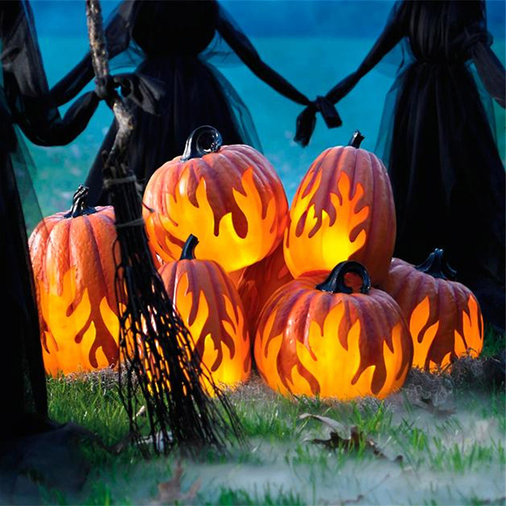 Beleuchtete orangefarbene Kürbisse mit Flammenausschnitten auf Gras, ein Besen und Silhouettenfiguren im Hintergrund, Halloween-Thema.