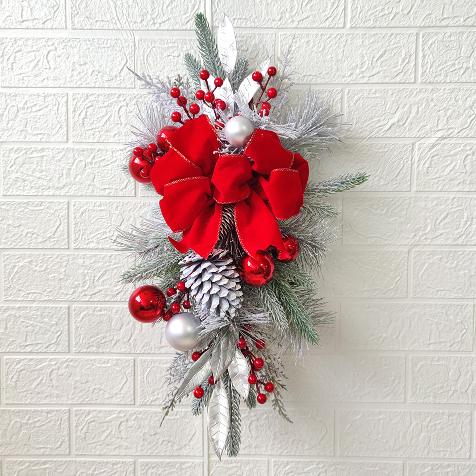 Eine Weihnachtsdekoration an einer weißen Ziegelmauer. Sie hat eine rote Schleife, Ornamente, Tannenzapfen und silberne Blätter.