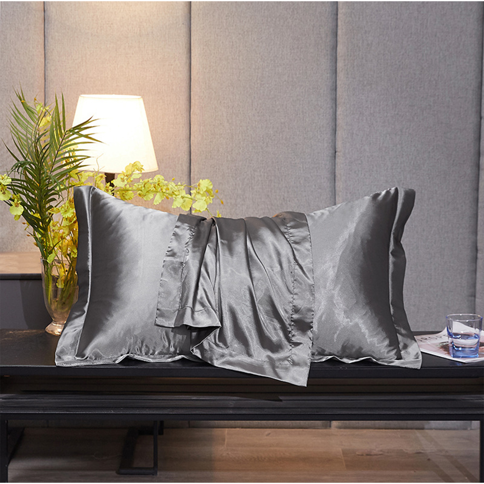 Ein graues Seidenkissen und ein gefaltetes Laken liegen auf einem schwarzen Tisch. Eine Lampe und Blumen sind hinter dem Kissen.