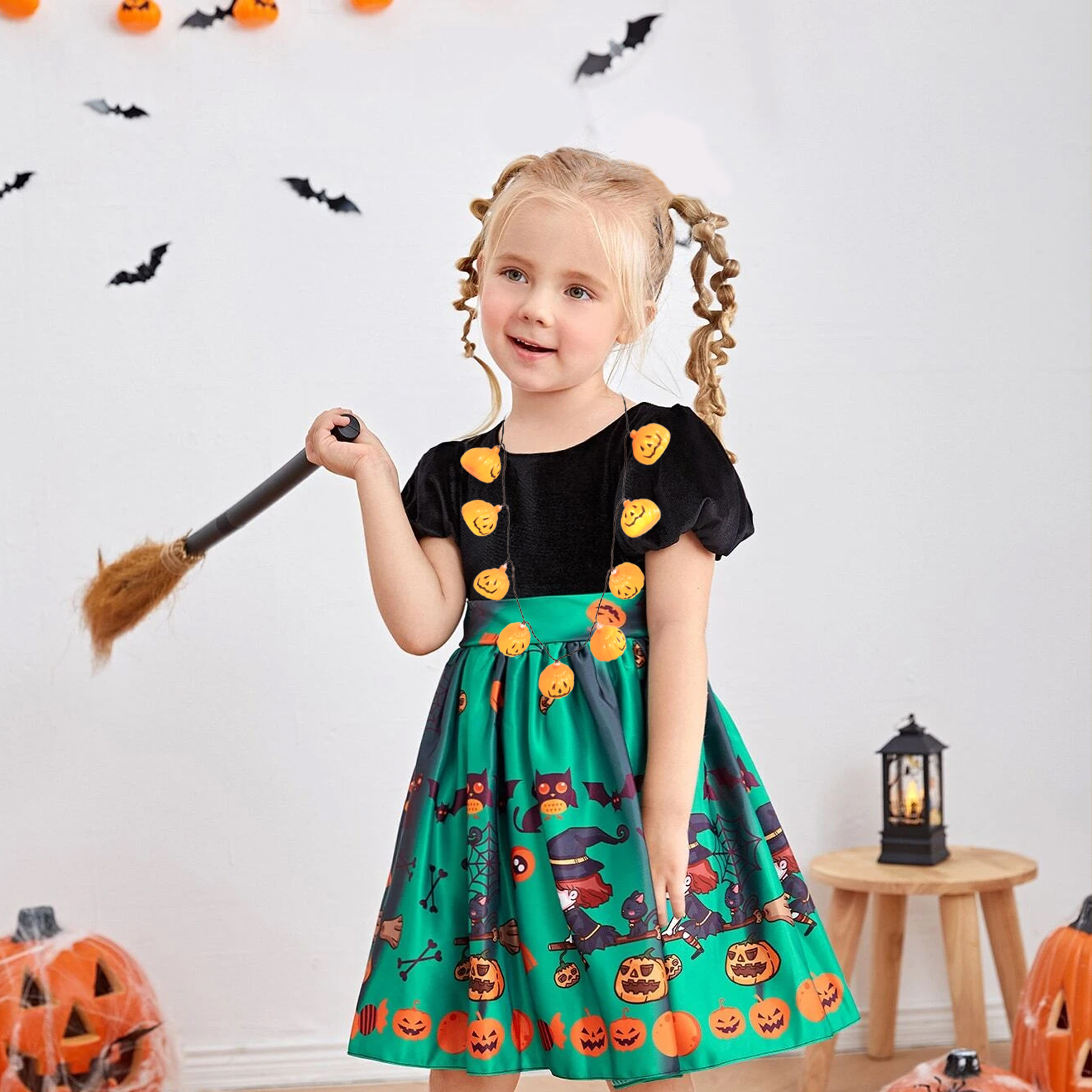 Ein Mädchen in einem Halloween-Kostüm hält einen Besen. Das Kleid ist grün mit Kürbissen und schwarzen Katzen.