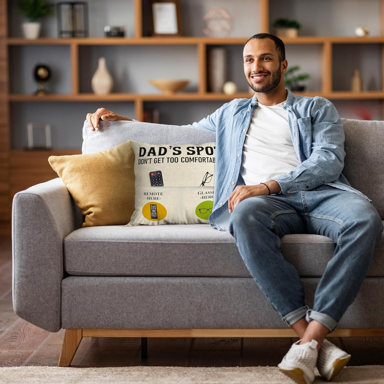Ein lächelnder Mann in Jeans sitzt auf einer Couch mit einem Kissen mit der Aufschrift 'Dad's Spot'. Im Hintergrund befindet sich ein Holzregal.