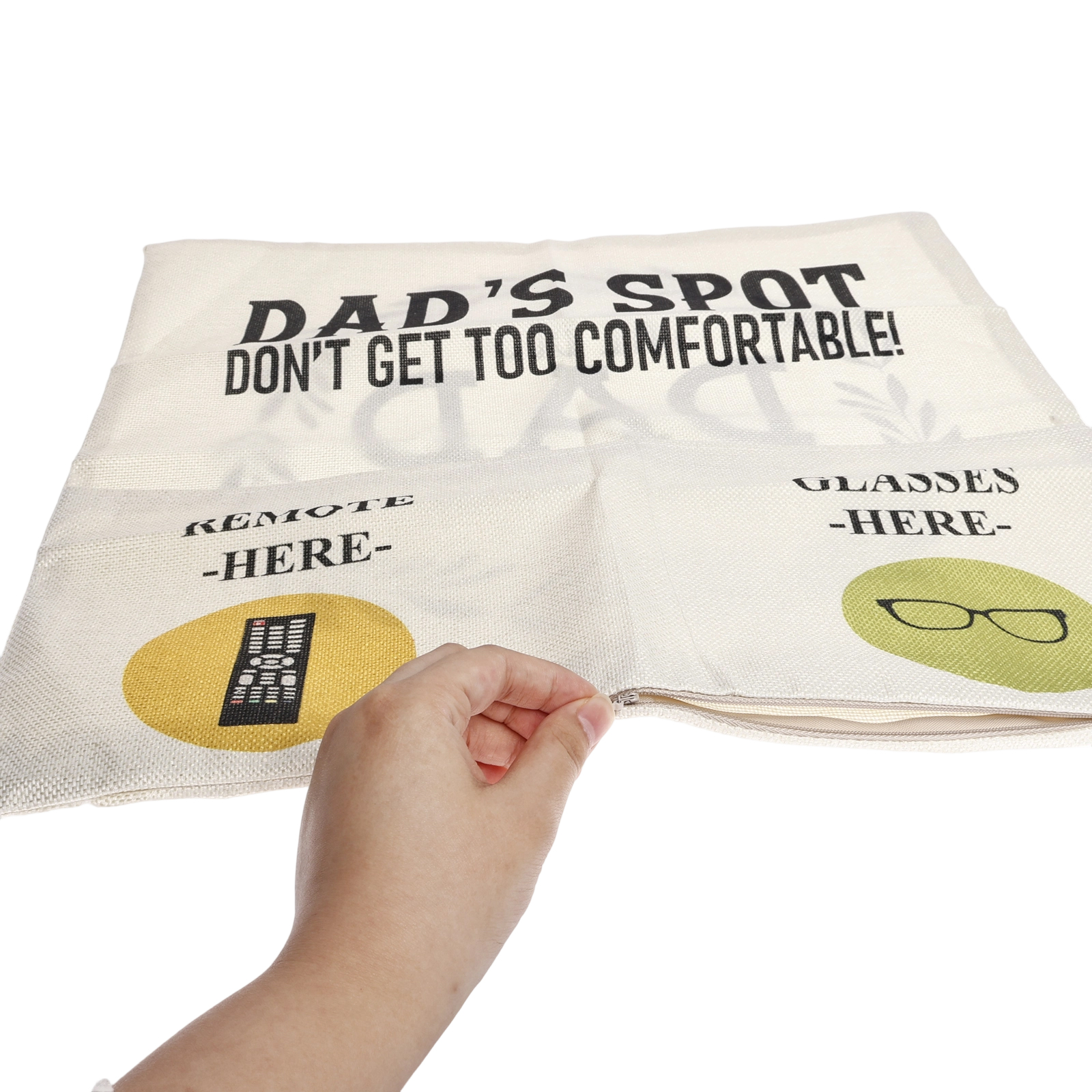 Eine Hand reißt einen Kissenbezug auf. Das Kissen hat Text: 'DAD'S SPOT DON'T GET TOO COMFORTABLE!' und 'REMOTE -HERE-'. 