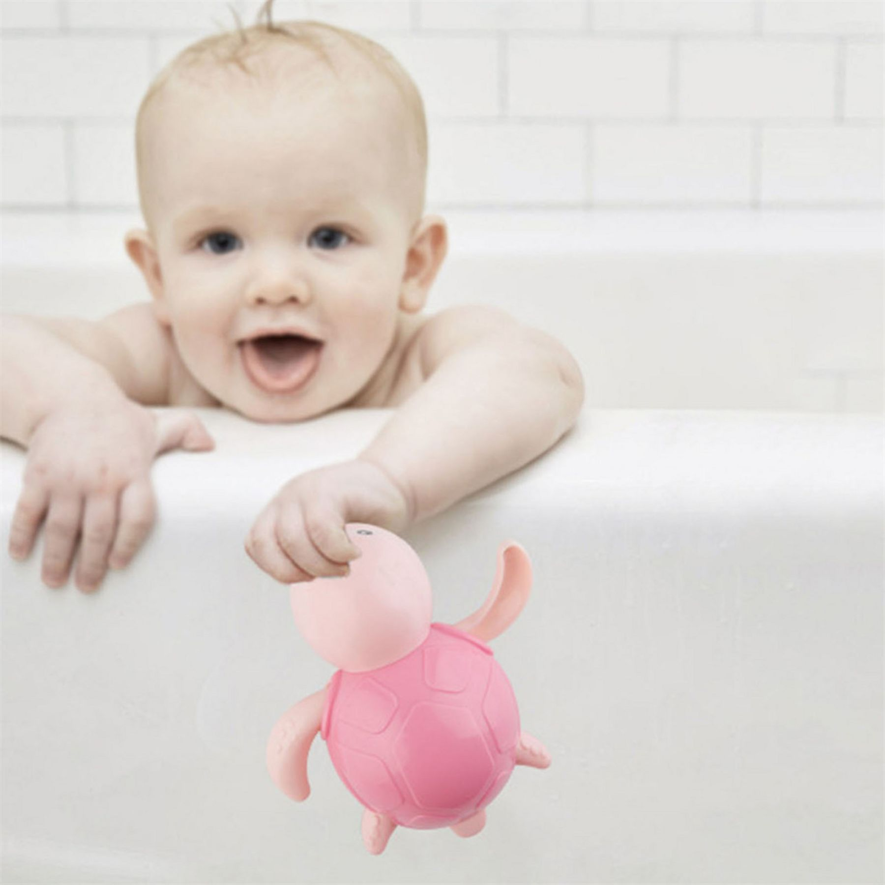 Ein Baby in einer Badewanne hält ein rosa Schildkrötenspielzeug. Das Baby lächelt mit offenem Mund. Weiße Kachelwand im Hintergrund.