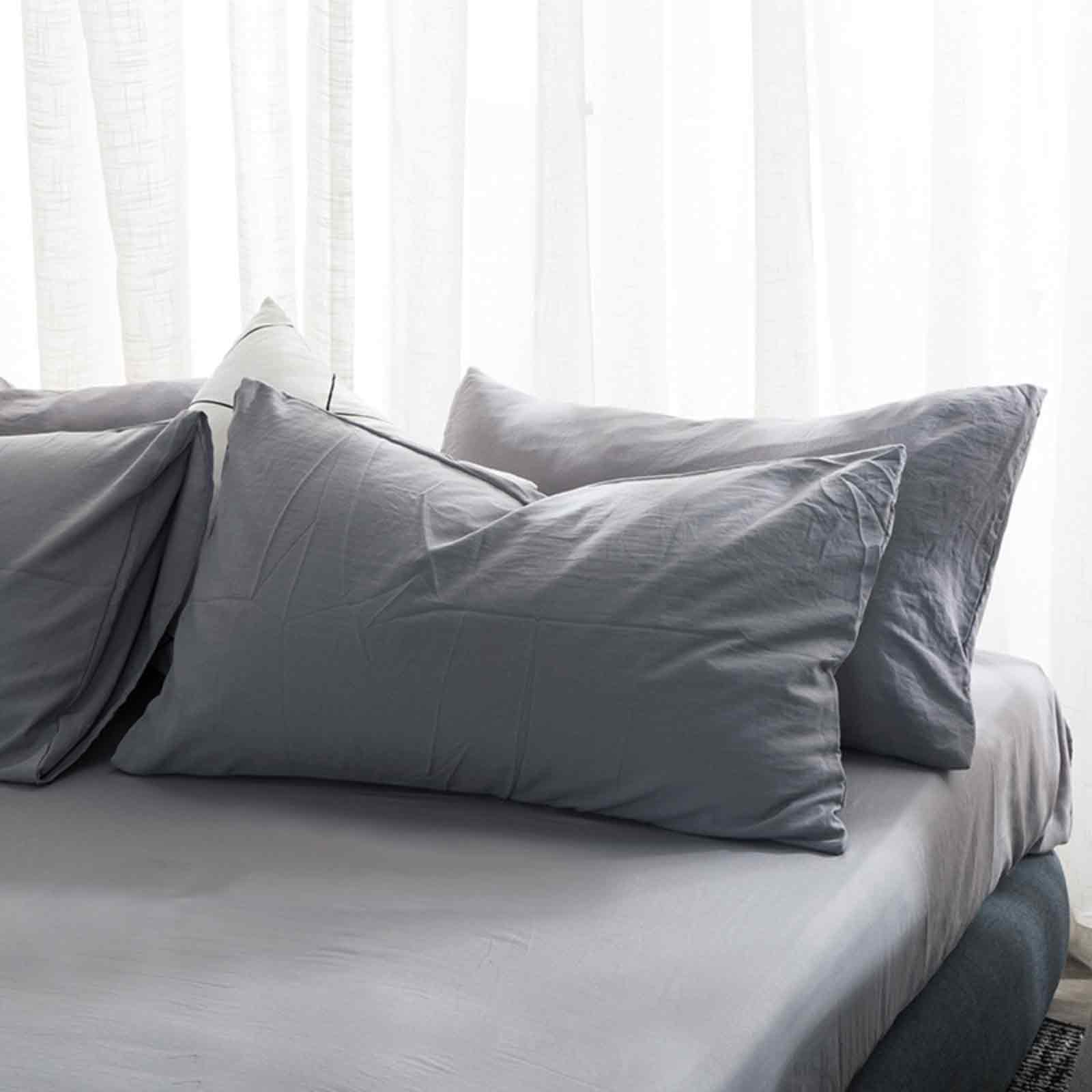 Bett mit grauer Bettwäsche und Kissen. Weiße Vorhänge im Hintergrund. Weiches Licht.
