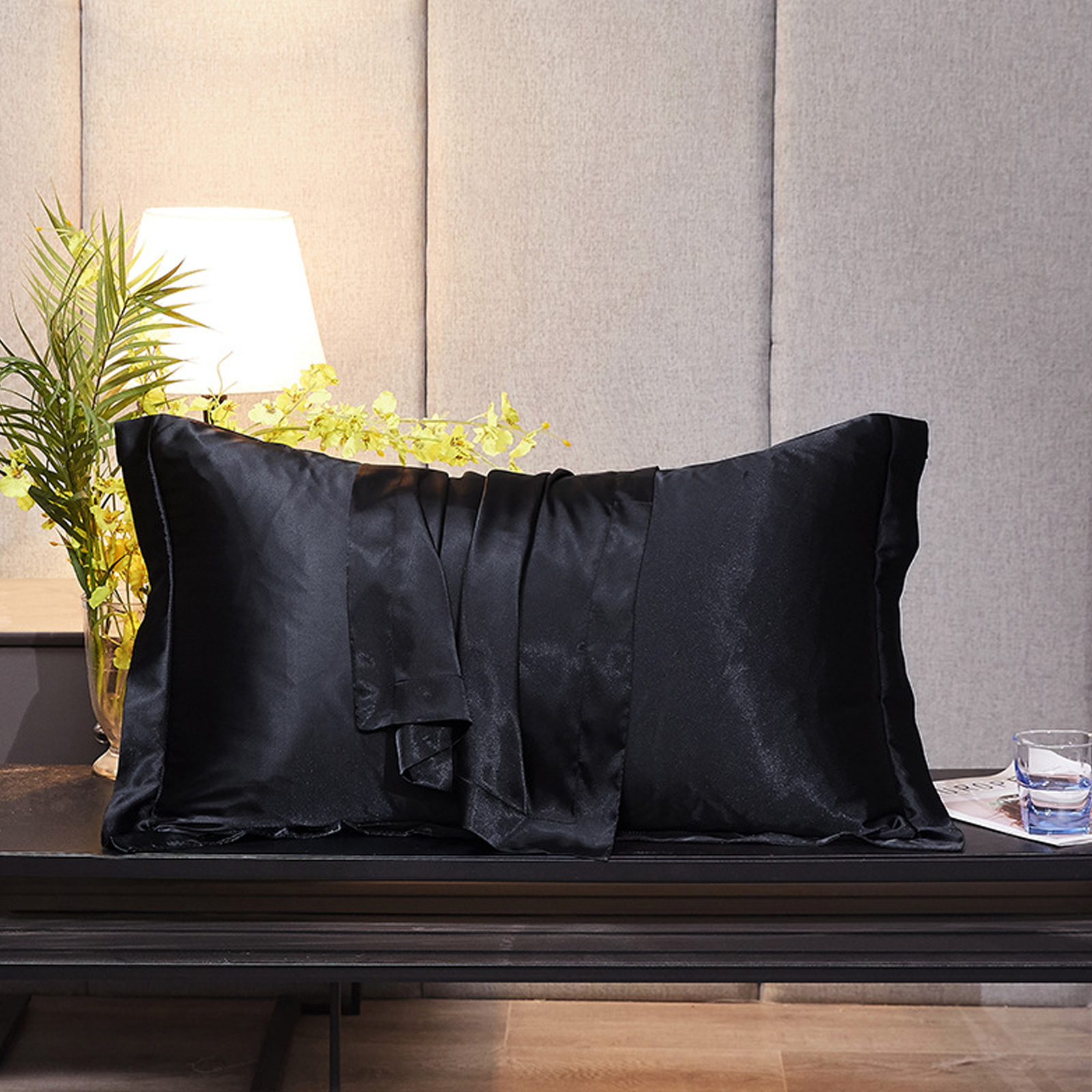 Ein schwarzes Satin-Kissen auf einem schwarzen Tisch. Eine Lampe, ein Glas Wasser und eine Pflanze befinden sich daneben, neutraler Hintergrund.