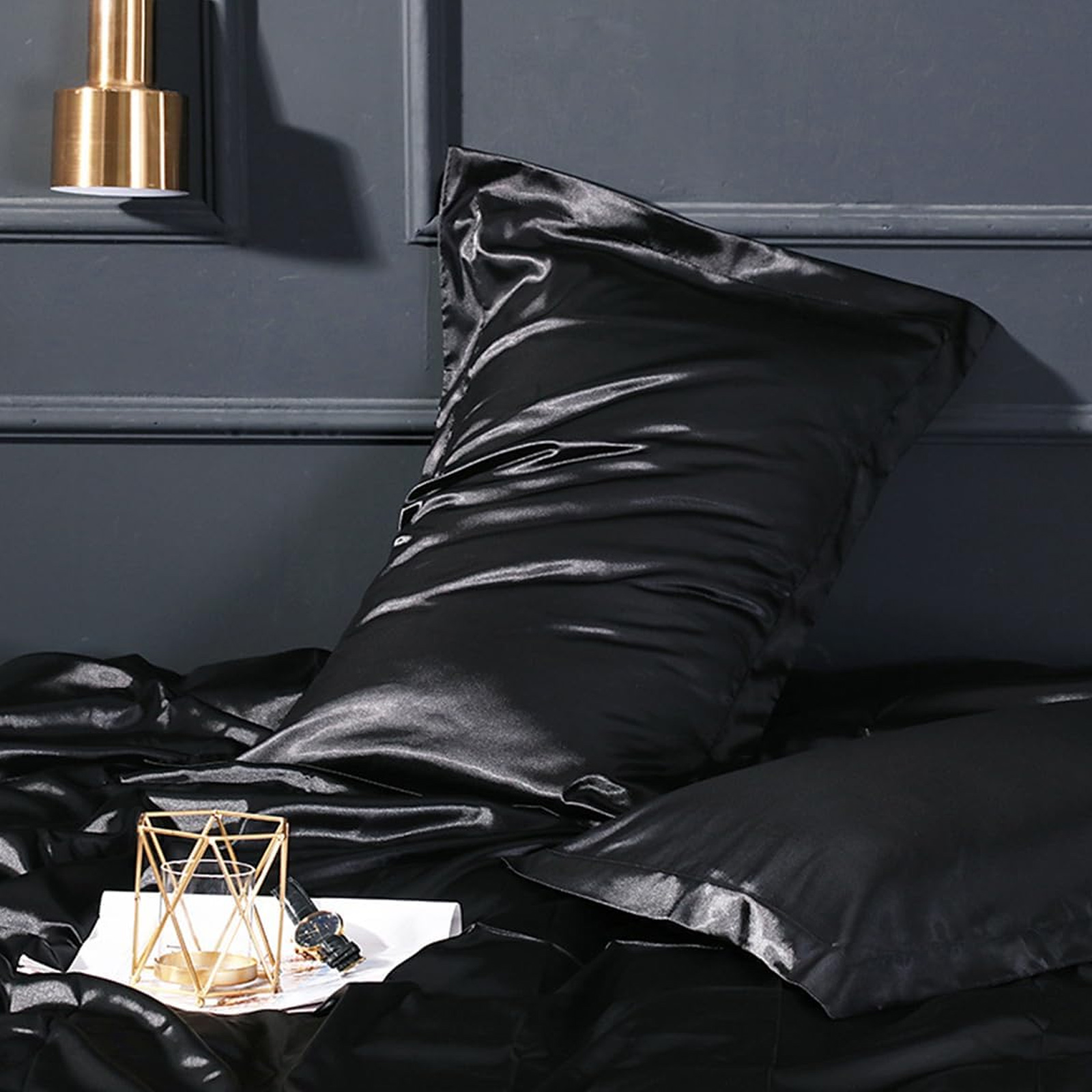 Ein schwarzes Satin-Kissen auf einem Bett mit einem goldenen geometrischen Kerzenhalter. Eine goldene Lampe hängt an der Wand.