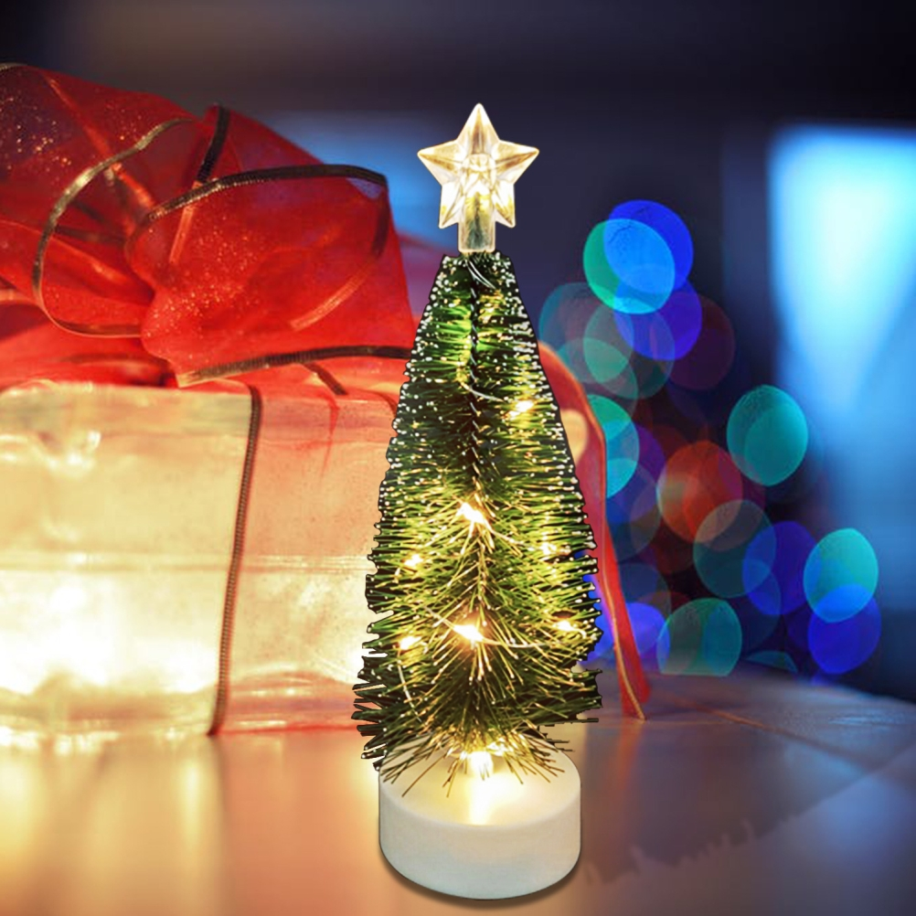 Ein kleiner, beleuchteter Weihnachtsbaum mit Stern. Daneben eine Geschenkbox mit roter Schleife, im Hintergrund verschwommene Lichter.