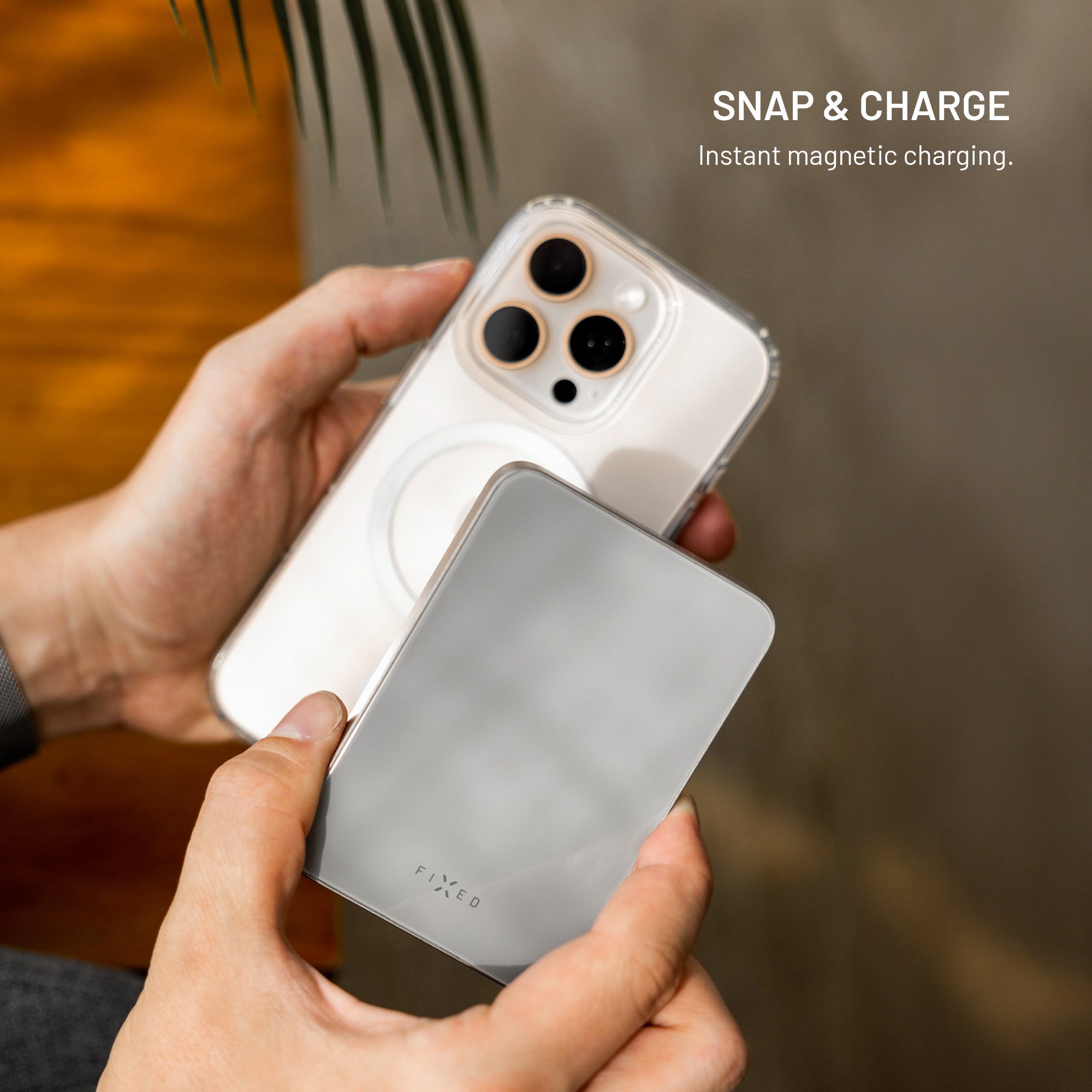 Hände halten ein Telefon mit einer transparenten Hülle und einem silbernen magnetischen Ladegerät vor unscharfem Hintergrund.