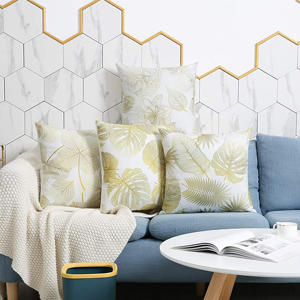 Ein blaues Sofa mit vier dekorativen Kissen mit goldenen Blättern. Eine weiße Decke, Tisch und Mülleimer sind im Bild.