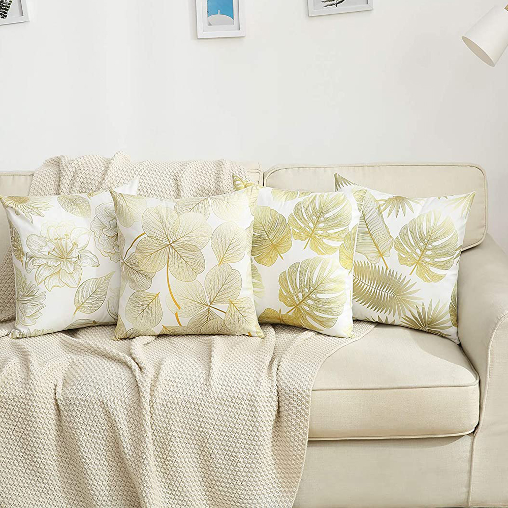 Vier dekorative Kissen auf einem beigen Sofa mit einer cremefarbenen Decke. Kissen haben goldene Blattmuster auf Weiß.