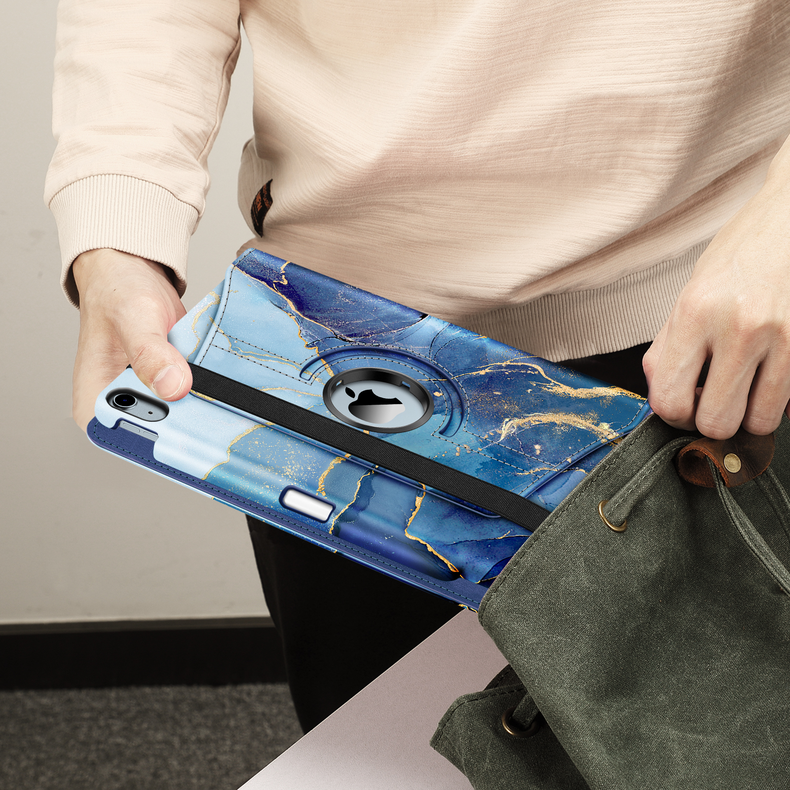 Person hält eine blaue Tablethülle mit Marmormuster und legt sie in eine grüne Canvas-Tasche. Die Person trägt einen beigen Pullover.