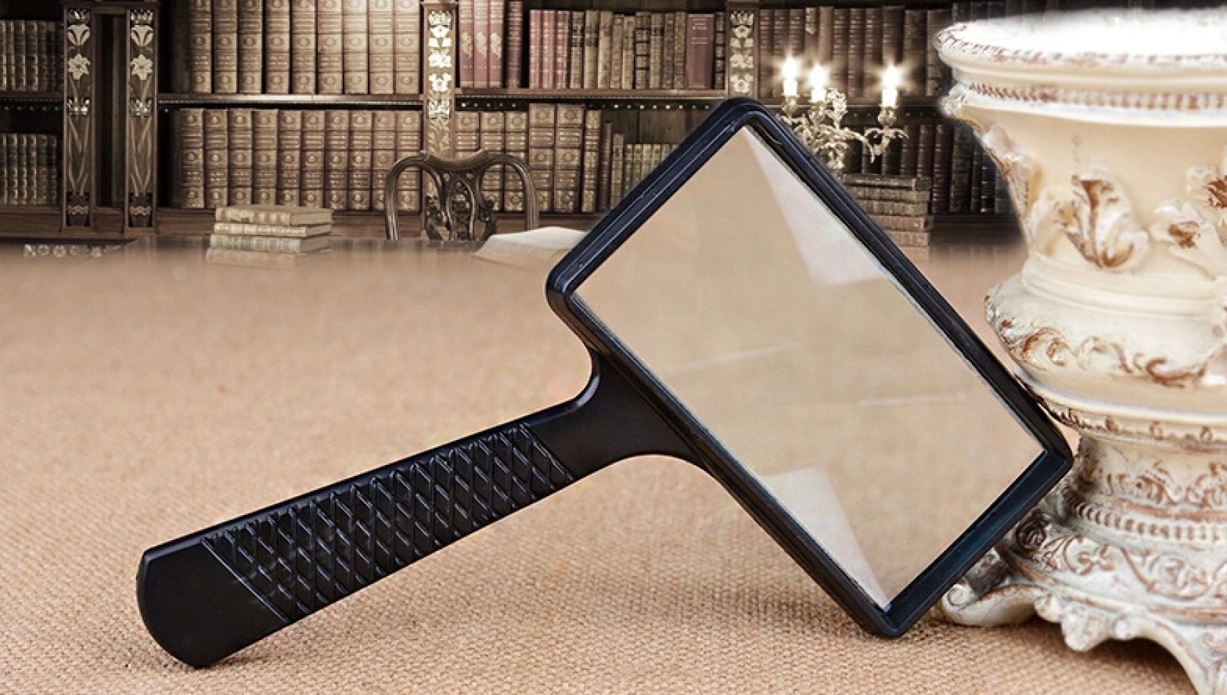 Eine schwarze Lupe liegt auf einer beigefarbenen Oberfläche. Im Hintergrund sind Bücherregale, Kerzen und eine Vase.
