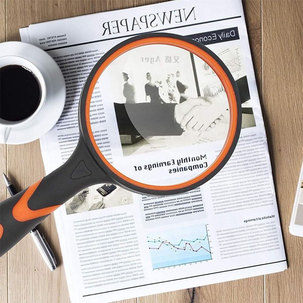 Eine Lupe über einer Zeitung. Eine Tasse Kaffee, ein Stift und ein Telefon liegen auf dem Holztisch.