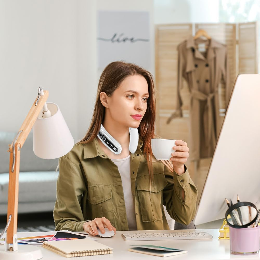 Eine Frau mit Nackenmassagegerät sitzt am Schreibtisch, hält eine Tasse und benutzt einen Computer. Eine Lampe, Tastatur und ein Mantel sind im Hintergrund.