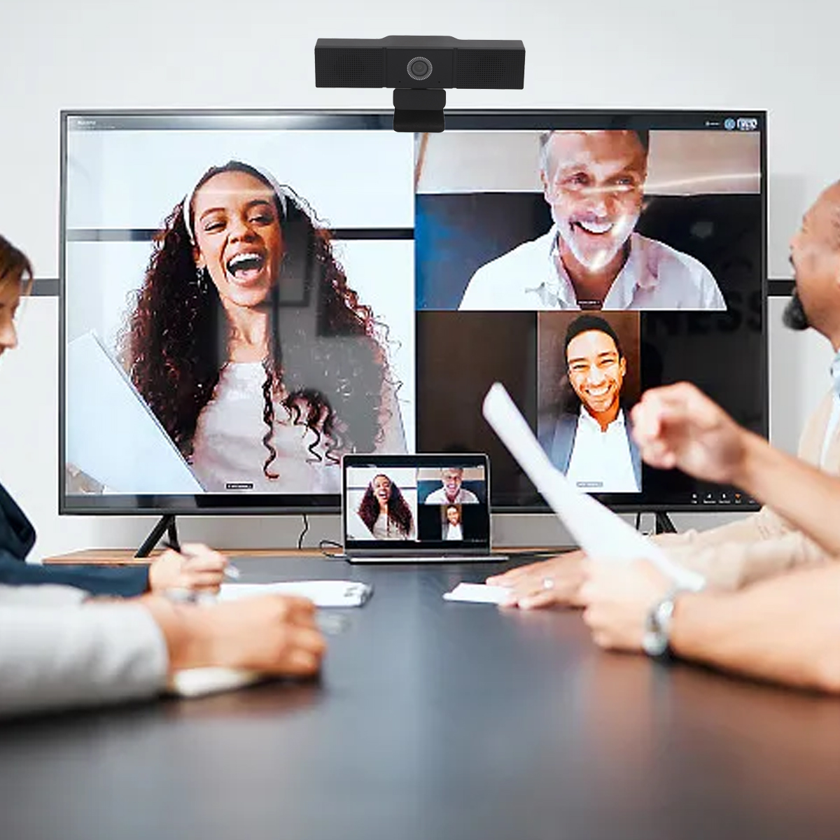 Besprechung im Gange. Personen sind in einem Videoanruf. Webcam über dem großen Bildschirm, Personen sitzen an einem Tisch.