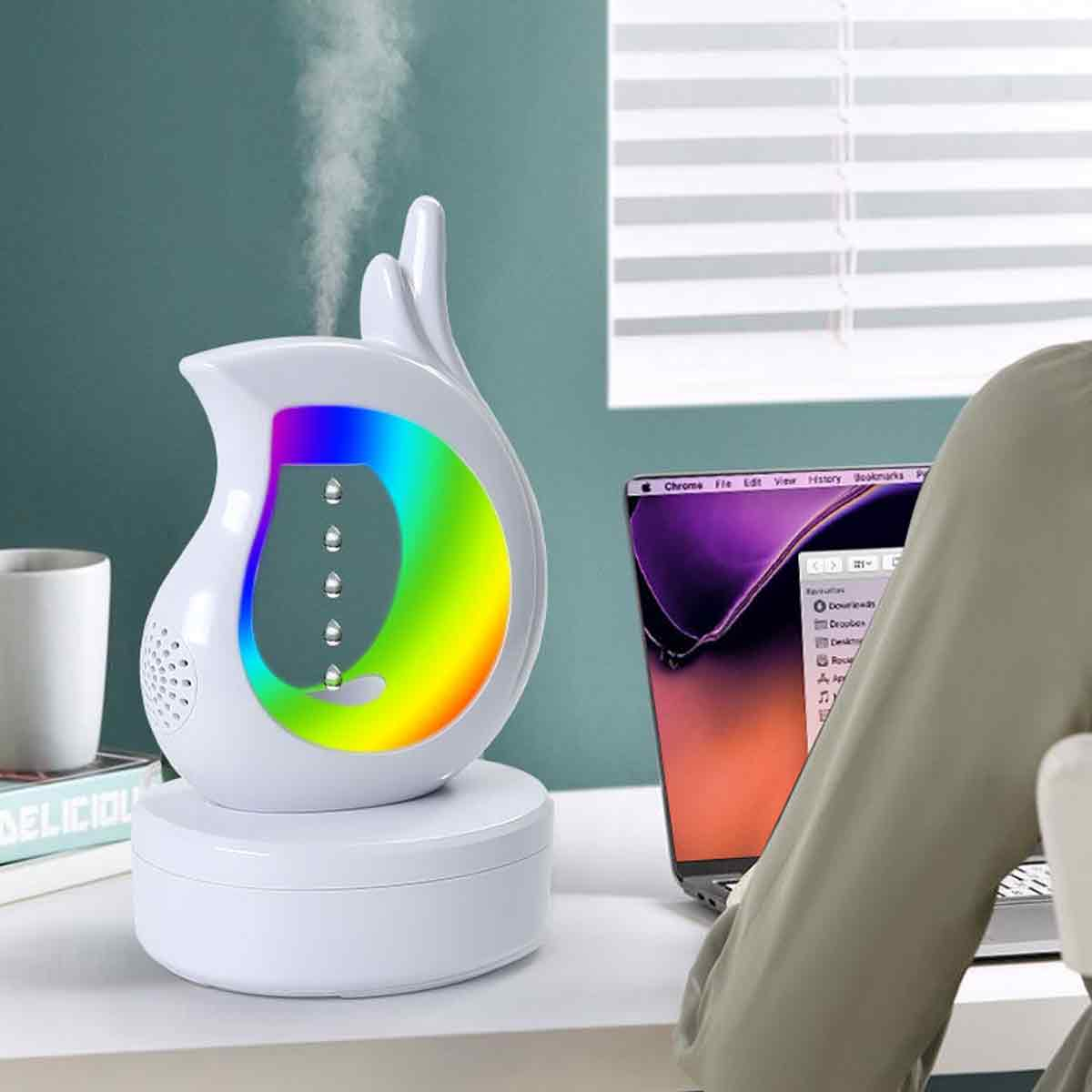 Ein weißer Luftbefeuchter mit Regenbogenlicht. Ein Laptop und eine Tasse stehen auf dem weißen Tisch. Person im Hintergrund.