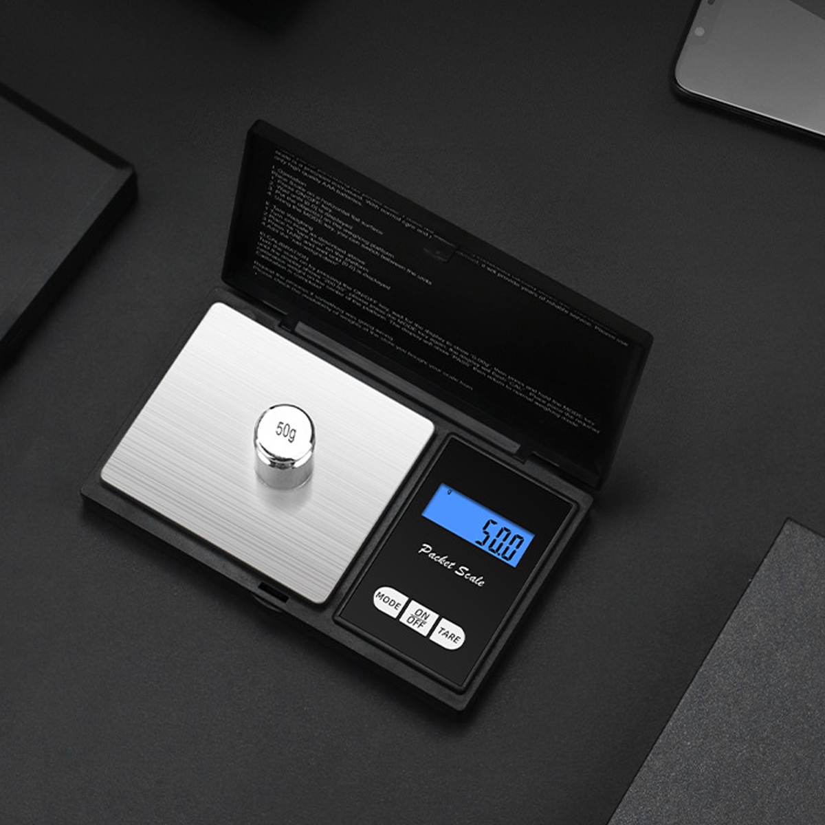 Eine Taschenwaage zeigt 50,0 an, mit einem 50g-Gewicht auf der Waage. Schwarzes Gehäuse mit Anweisungen und ein Telefon im Hintergrund.