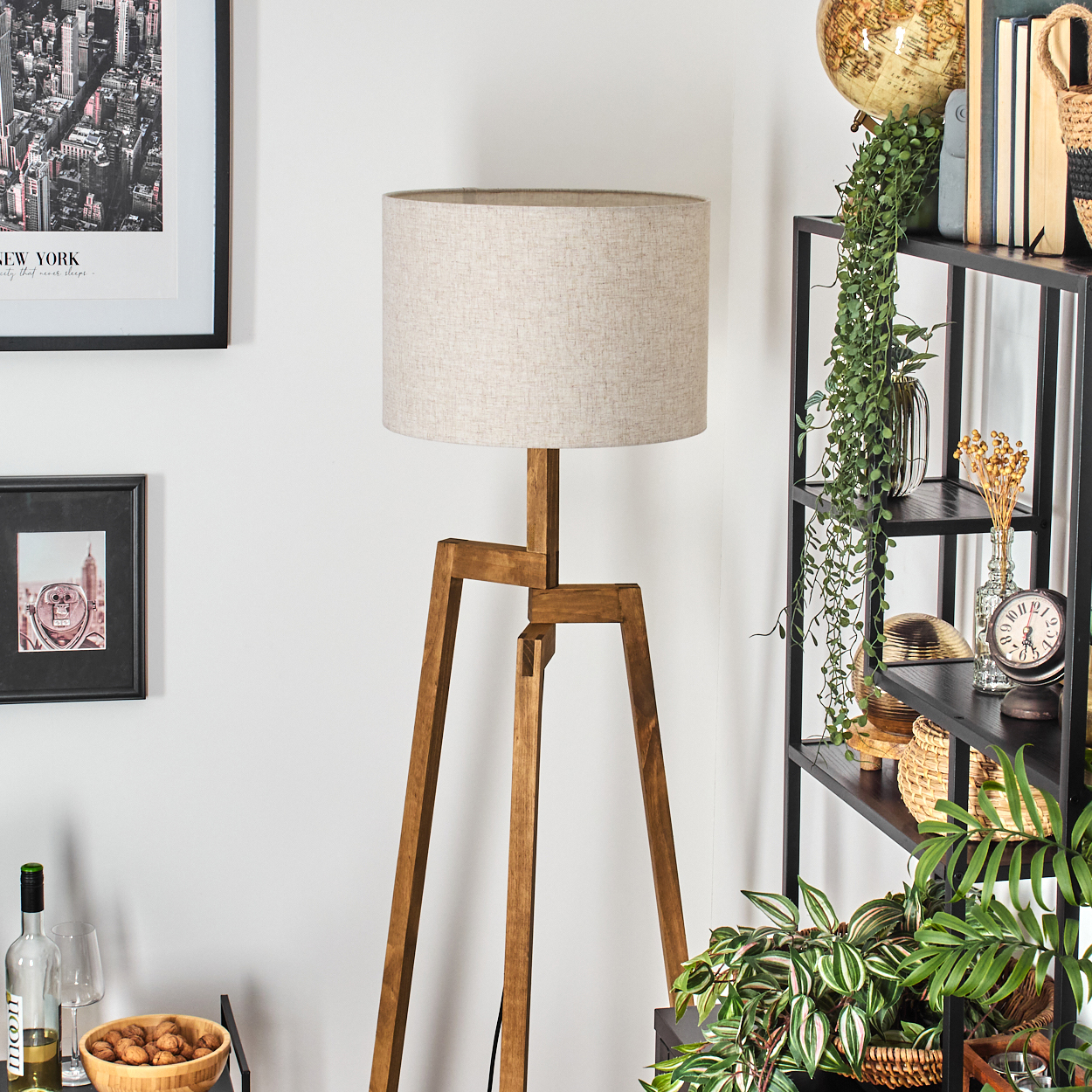 Eine hölzerne Stativ-Stehlampe mit beigem Schirm steht in einem Raum. Regale mit Pflanzen und Dekorationen sind im Hintergrund.