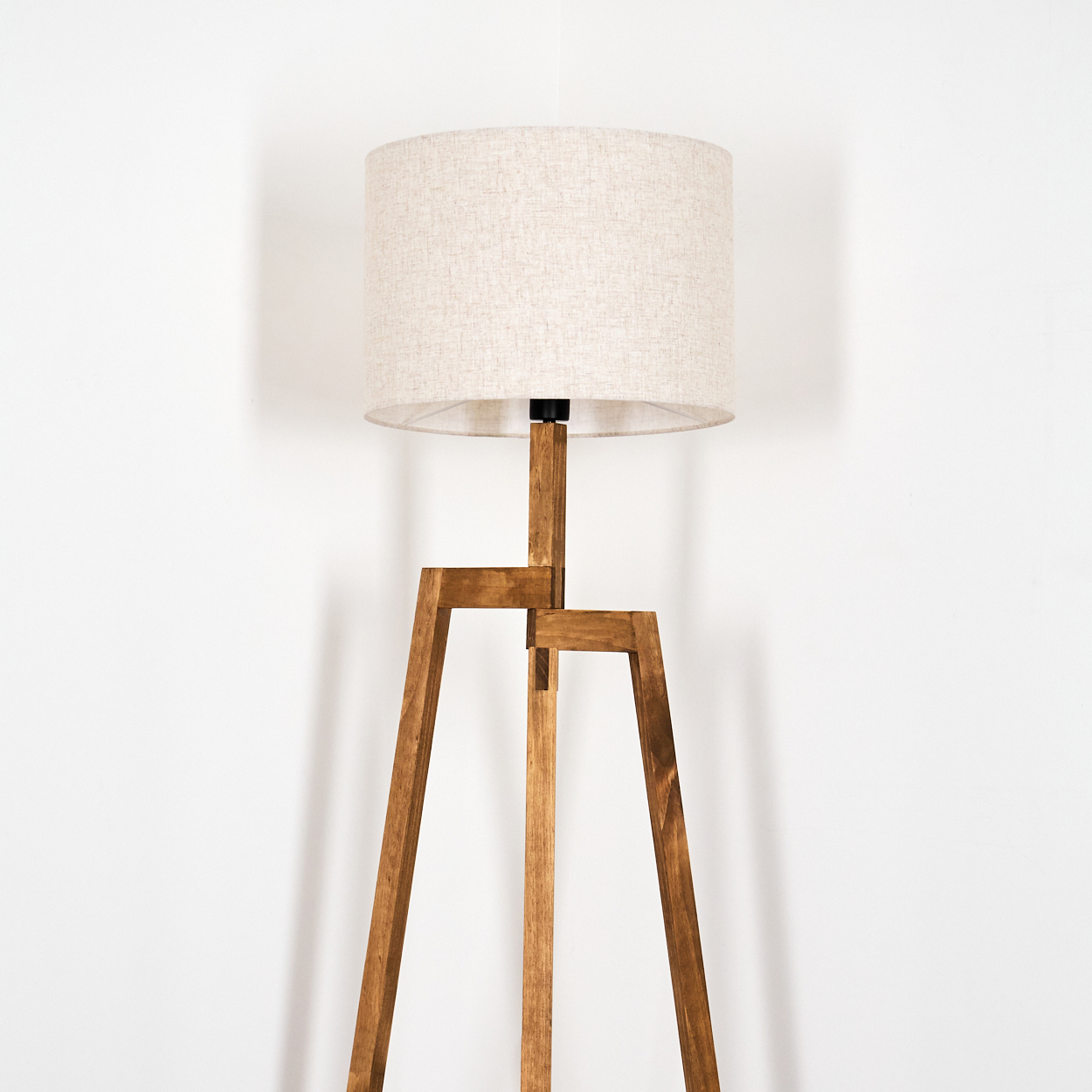 Eine hölzerne Stehlampe mit beigem Lampenschirm steht vor einer weißen Wand. Die Lampe hat ein schlichtes, modernes Design.