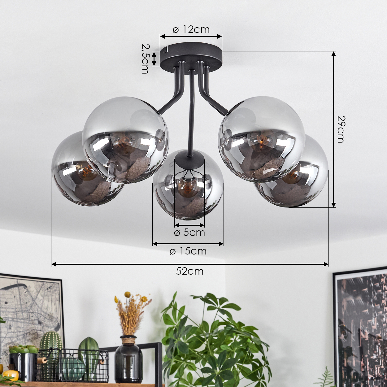 Eine moderne Deckenlampe mit fünf kugelförmigen Schirmen. Die Lampe ist schwarz, mit beschrifteten Maßen. Darunter ist ein Regal mit Pflanzen zu sehen.