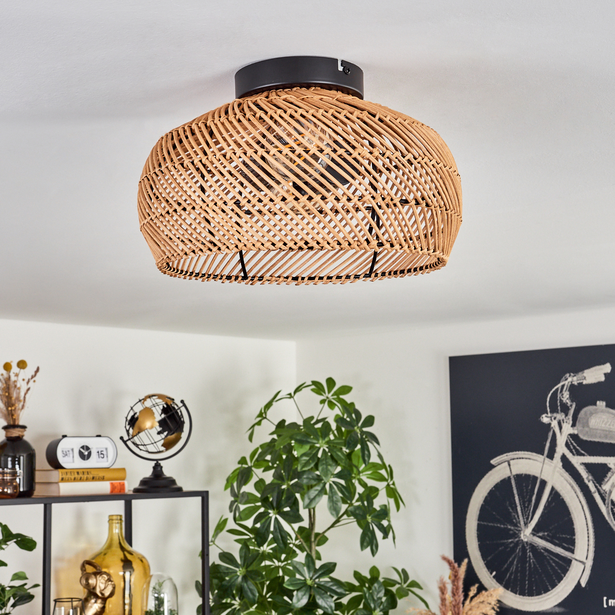 Eine Deckenlampe aus geflochtenem Rattan ist das zentrale Objekt. Darunter ist ein Regal mit Dekoration zu sehen. Ein Fahrradbild hängt an der Wand.