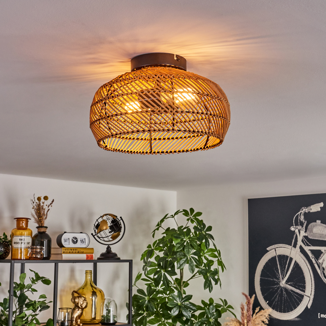 Gewebte Deckenlampe aus Rattan, die einen Raum beleuchtet. Darunter ein Regal mit Dekoration und einer Pflanze sowie ein Fahrradkunstwerk.