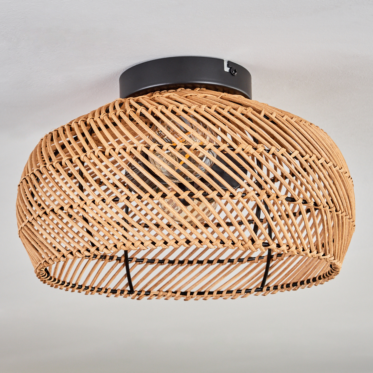 Ein gewebter Lampenschirm aus Rattan, braun, mit schwarzem Metallsockel, hängt von einer weißen Decke. Im Inneren ist eine Glühbirne sichtbar.