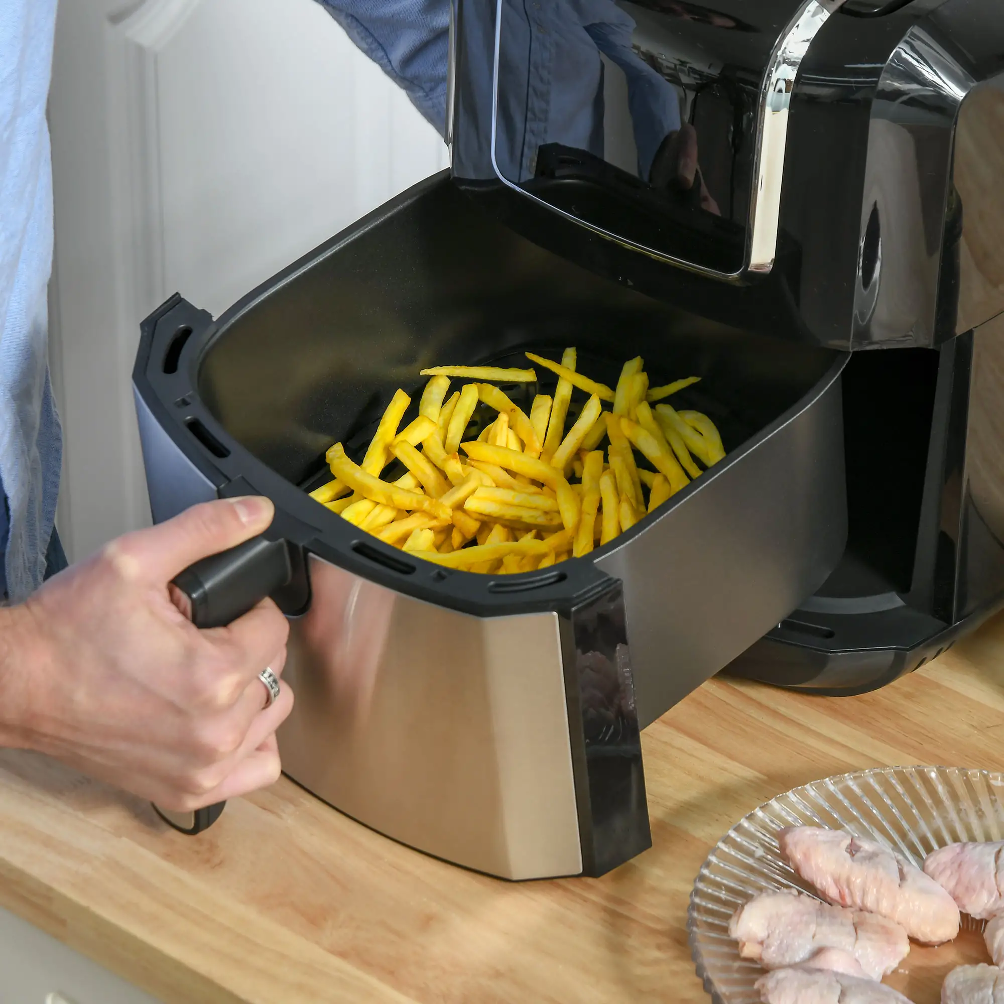 Ein Airfryer mit Pommes Frites, eine Person, die den Griff des Fryers hält, und ein Teller mit rohen Hähnchenflügeln.