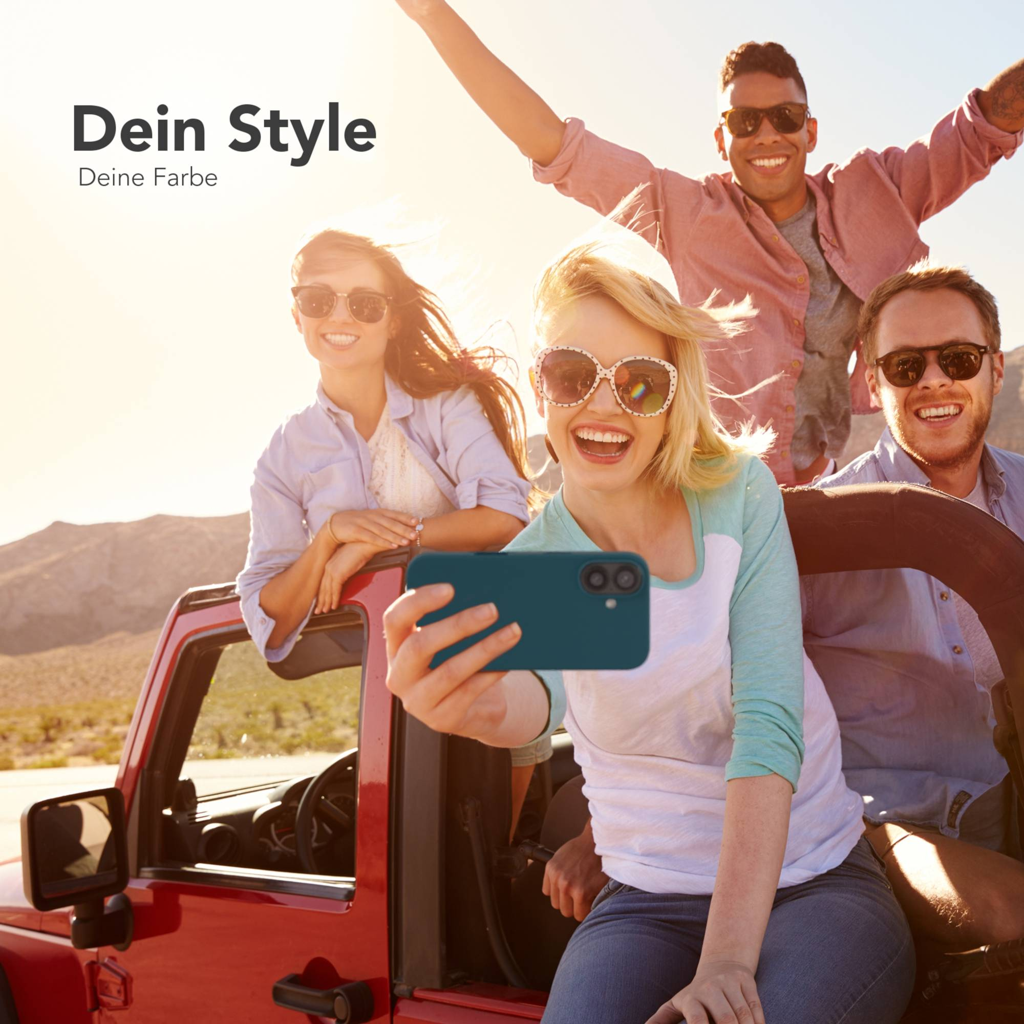 Vier Freunde in einem roten Jeep machen ein Selfie. Blonde Frau hält das Telefon. Sonniger Tag, Wüstenhintergrund.