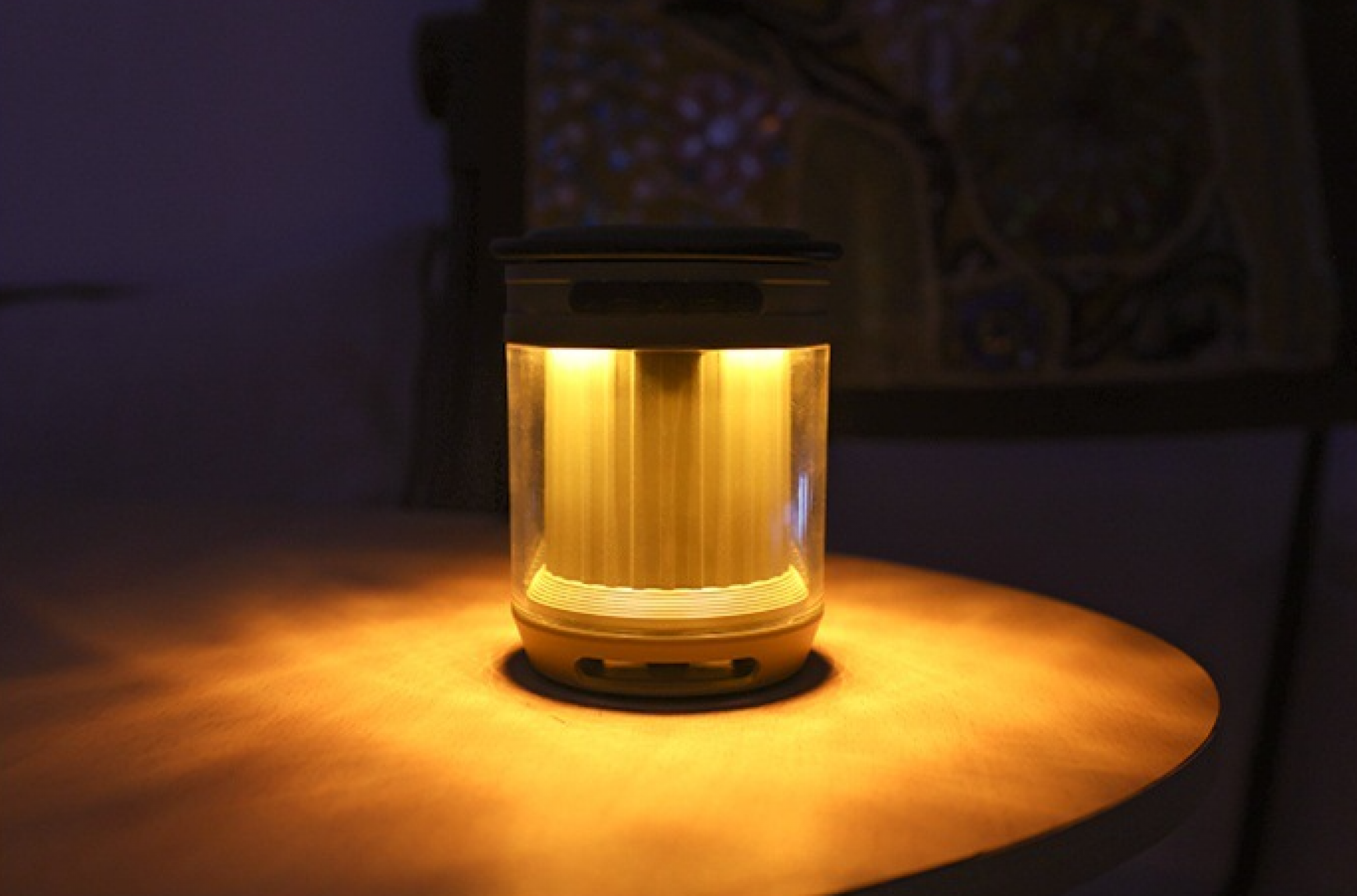Eine leuchtende zylindrische Lampe auf einem Holztisch. Der Hintergrund ist dunkel und ein Design an der Wand.