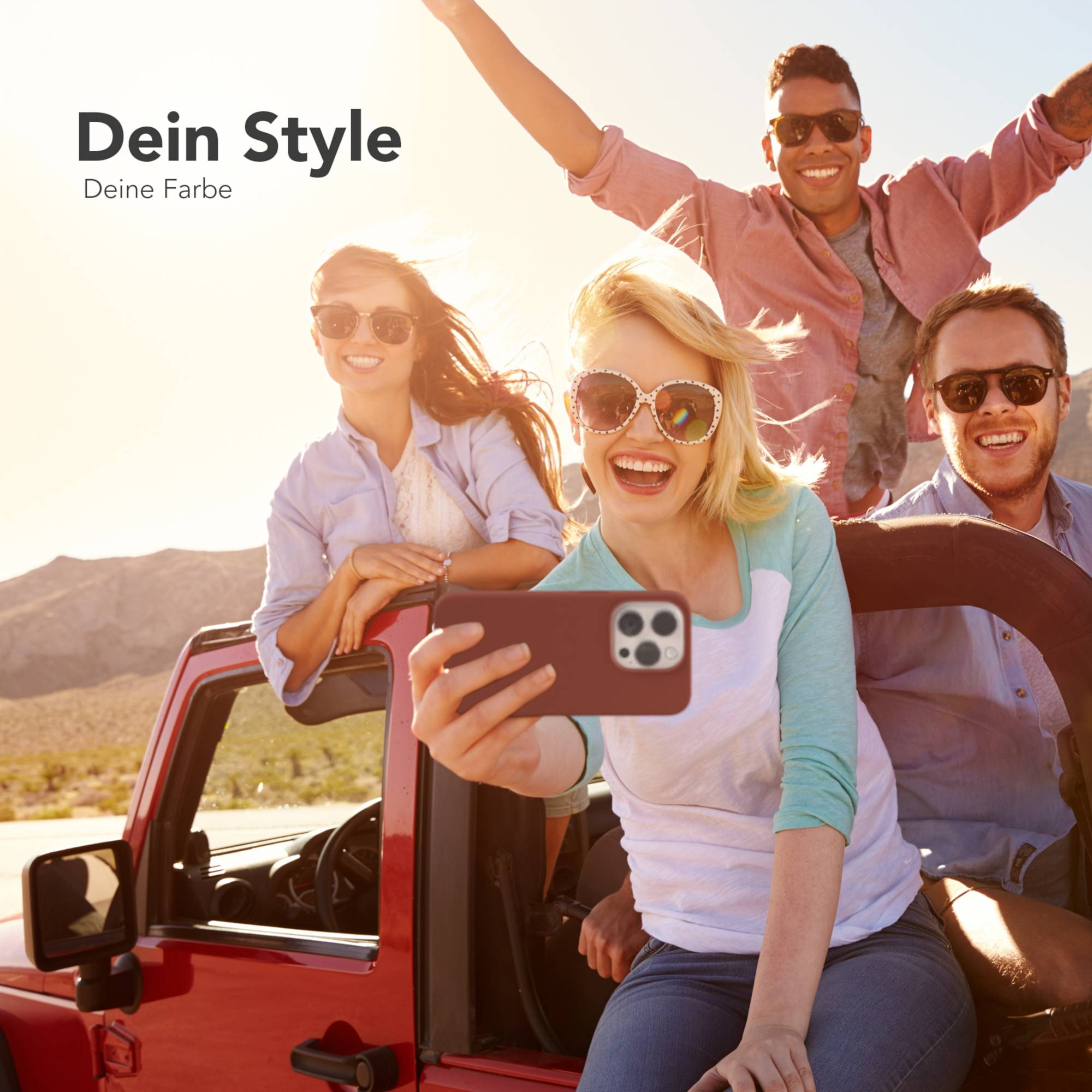 Vier lächelnde Personen mit Sonnenbrillen machen ein Selfie in einem roten Jeep vor einer Wüstenkulisse. Heller Sonnentag.