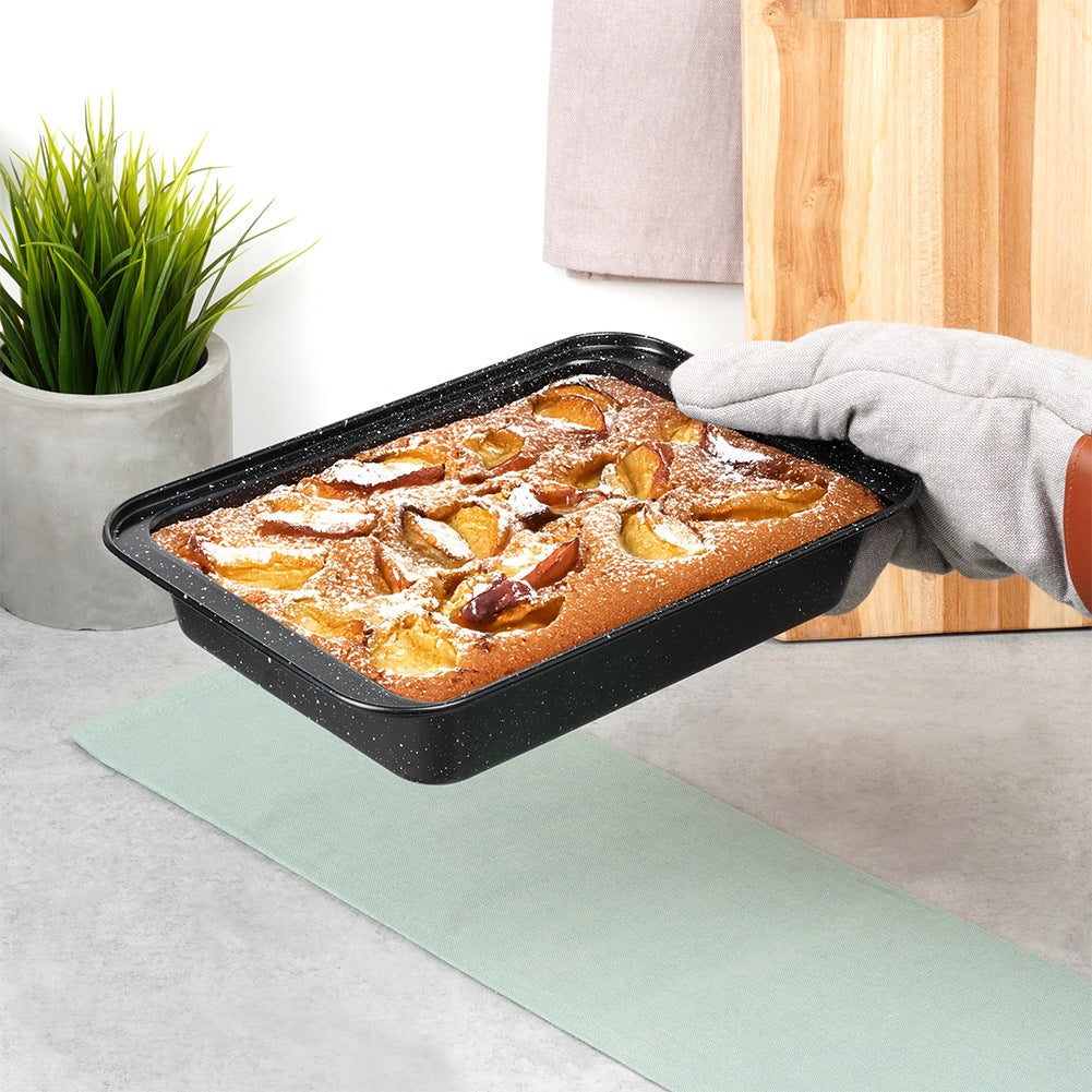 Una mano con un guante de horno sostiene una bandeja para hornear con un pastel cubierto de fruta. Una estera verde y una tabla de madera están en el fondo.