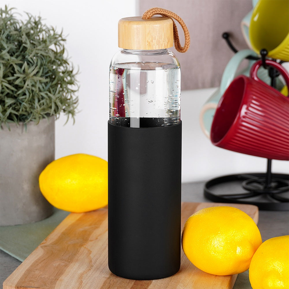 Una botella de agua de vidrio con tapa de madera y funda negra, limones y una planta sobre una tabla de madera. Tazas en el fondo.