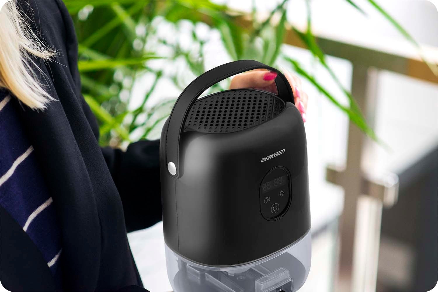 Una mujer sostiene un purificador de aire Berosen negro. Tiene un asa y una pantalla digital. El fondo es follaje verde borroso.